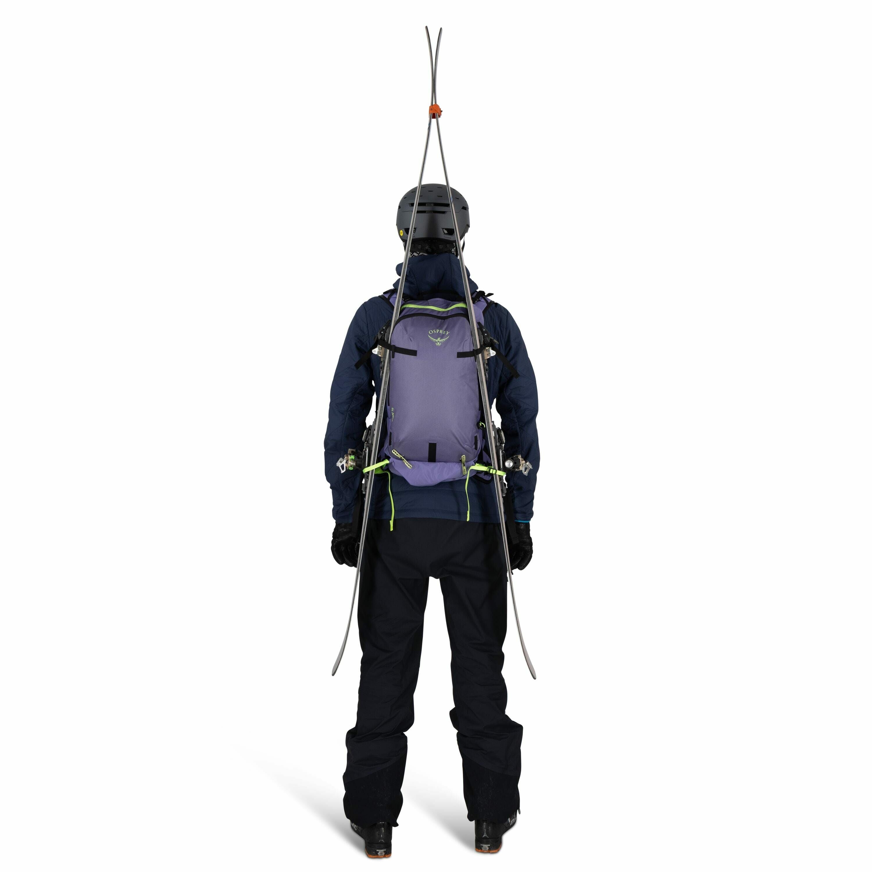 The image shows a person viewed from the back, equipped for skiing or mountaineering. They are wearing a helmet, a dark jacket, and dark pants, with a pair of skis fastened upright to a light purple backpack. The backpack has multiple straps and fastening
