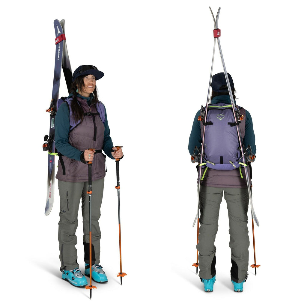 The image shows an individual dressed in outdoor skiing gear, standing with skis attached to a backpack. The person is wearing a purple and blue jacket layered over other clothing, with dark gray pants and blue ski boots. They are holding ski poles in bot