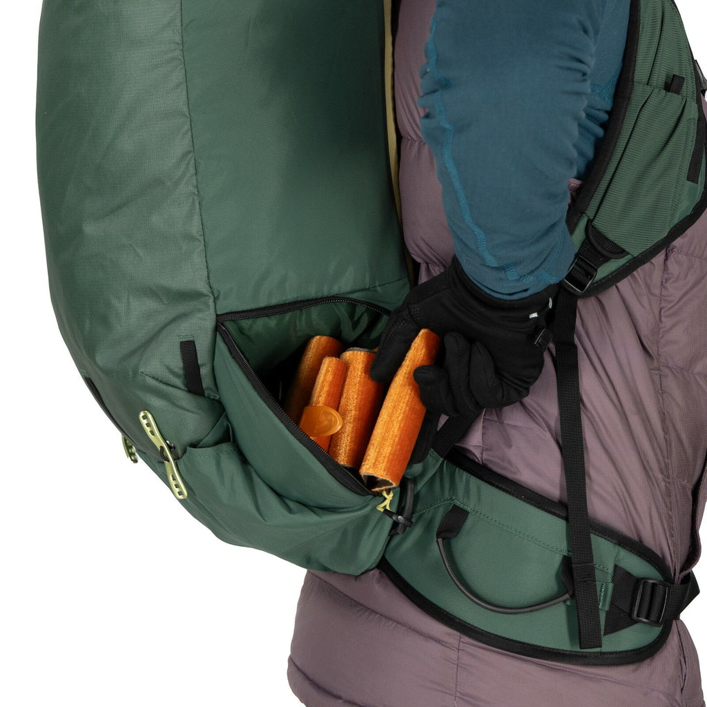 The image shows a person wearing a large green backpack with a side zipper pocket. The pocket is partially open, revealing a group of orange trekking poles inside. The person is wearing outdoor clothing, including a teal and dark gray jacket with long sle