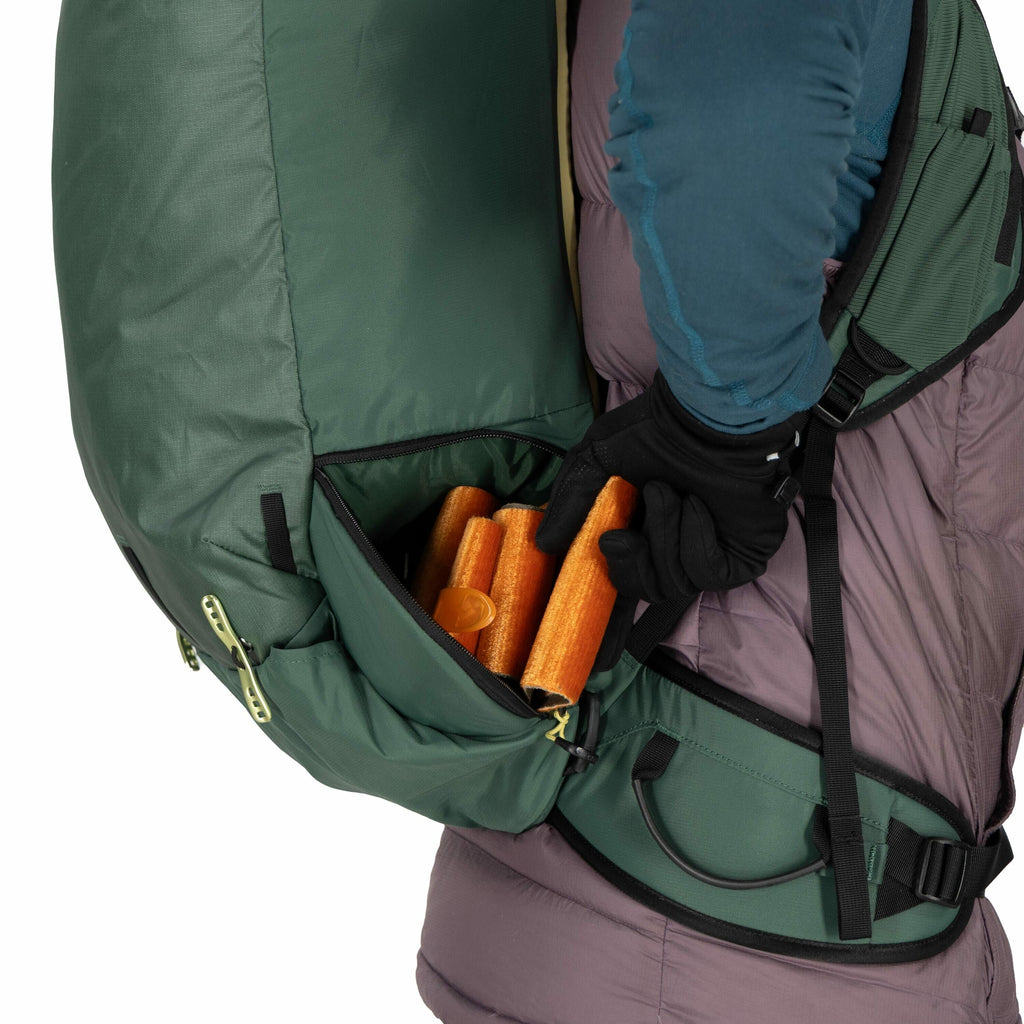 The image shows a person wearing a large green backpack with a side zipper pocket. The pocket is partially open, revealing a group of orange trekking poles inside. The person is wearing outdoor clothing, including a teal and dark gray jacket with long sle