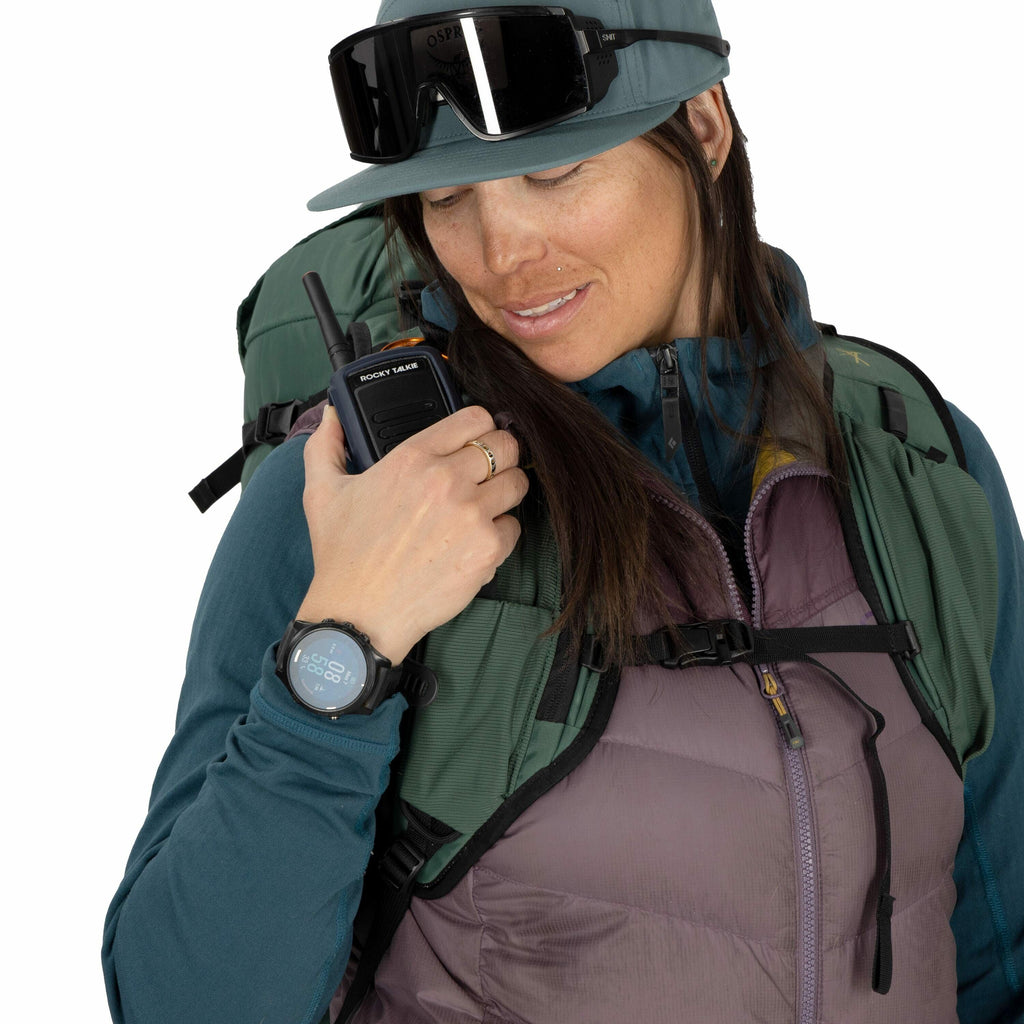 The image depicts a person holding a portable radio in their right hand, possibly communicating or checking the device. They are dressed for outdoor activities, wearing a teal-colored cap and a layered ensemble consisting of a teal long-sleeve shirt, a pu