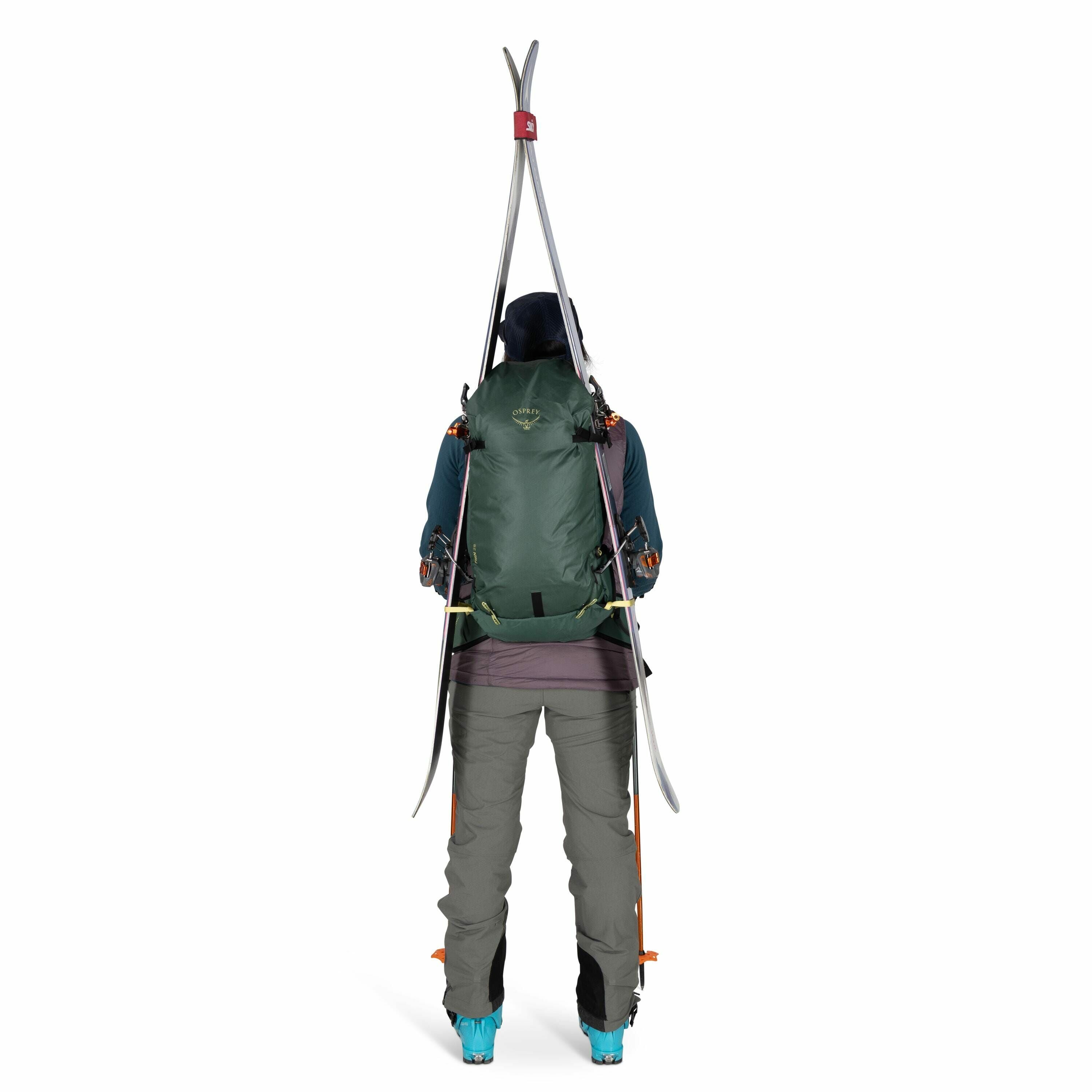 The image shows a person standing with their back to the camera, wearing a backpack and ski equipment. The individual is dressed in winter gear, including gray pants and a teal jacket, suitable for skiing. The backpack is dark green with orange accents an