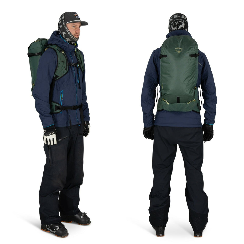 The image shows a person dressed in outdoor winter attire, viewed from both the front and back. They are wearing a navy blue jacket with a high collar and black pants suitable for cold weather activities. The individual is equipped with a large, green bac