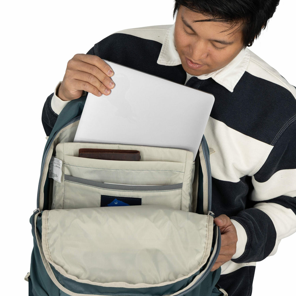 The image shows a person in a black and white striped long-sleeve shirt placing or removing a laptop from a backpack. The backpack is open, revealing an organized interior with various compartments, one of which holds the laptop. Additionally, there appea