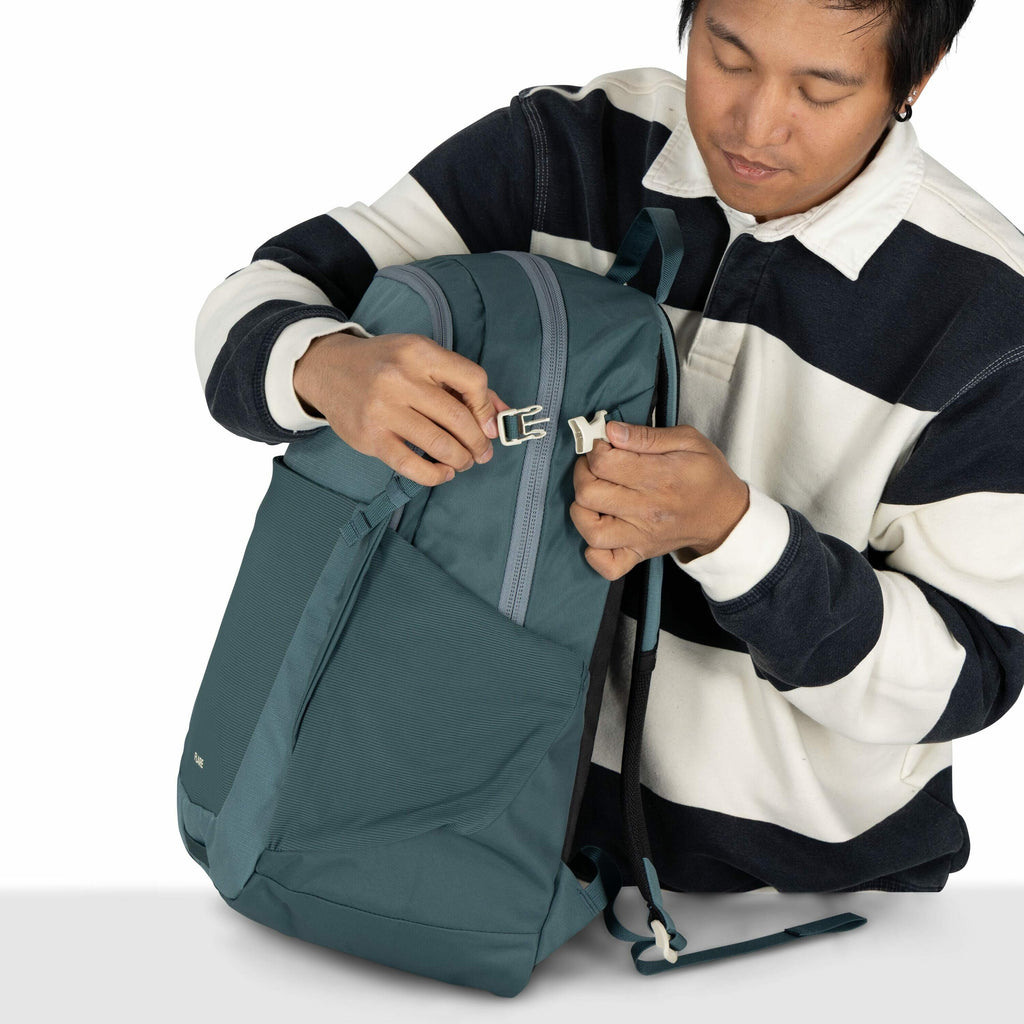 The image shows a person holding a teal-colored backpack, which they are in the process of securing by fastening a white buckle. The backpack has a sleek design with a large front pocket and a visible zipper on top. The person is wearing a black and white