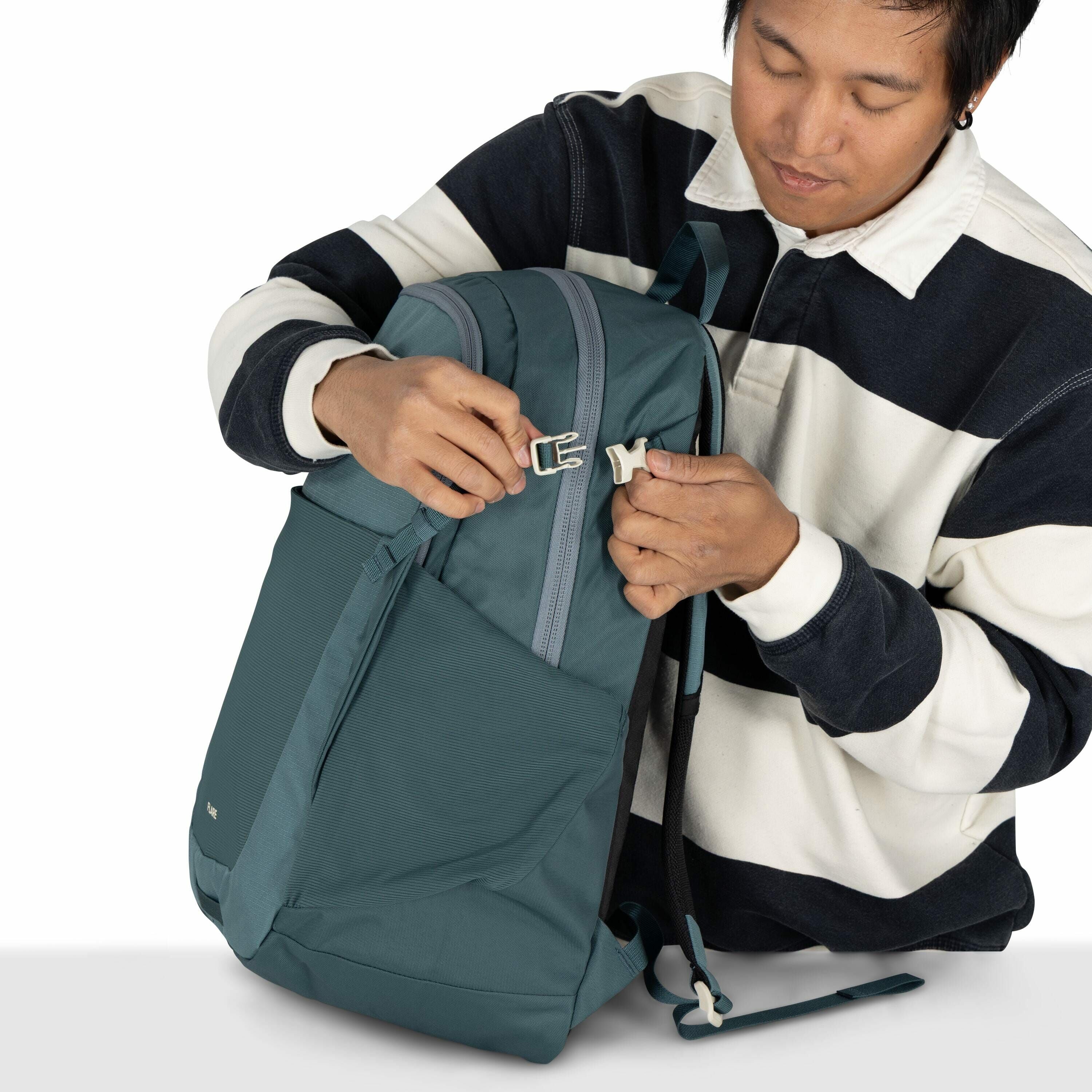 The image shows a person holding a teal-colored backpack, which they are in the process of securing by fastening a white buckle. The backpack has a sleek design with a large front pocket and a visible zipper on top. The person is wearing a black and white
