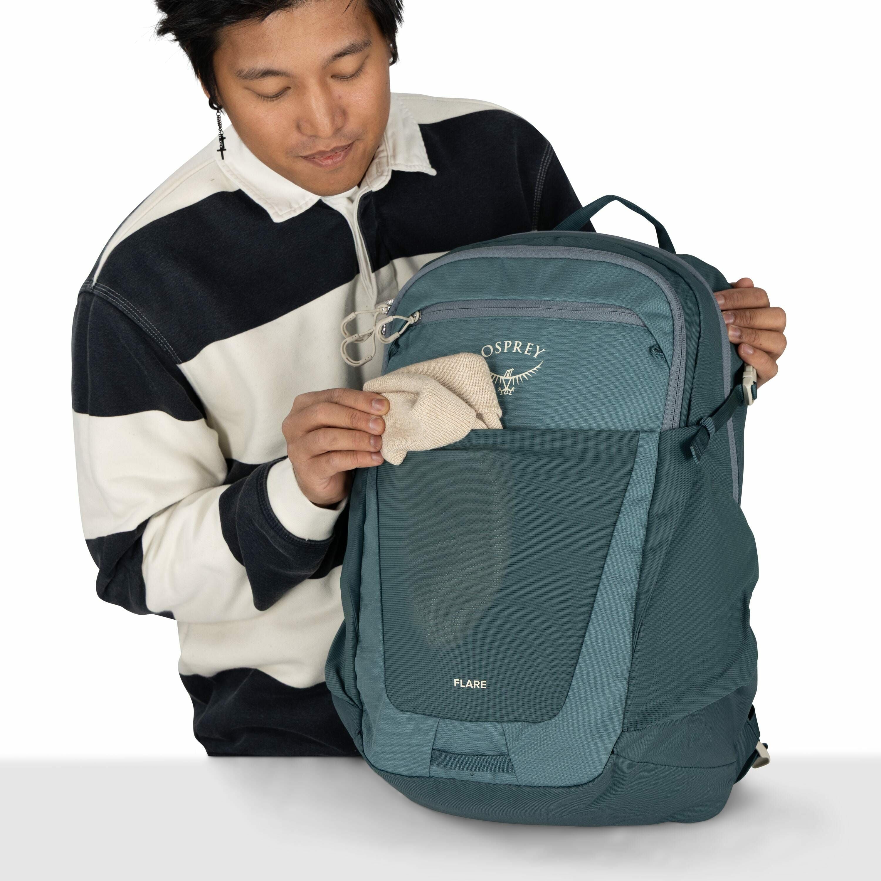 The image shows a person holding an Osprey Flare backpack. The backpack is teal in color and features multiple compartments and pockets. The person is placing a beige item, possibly a small towel or cloth, into the mesh pocket on the front of the backpack