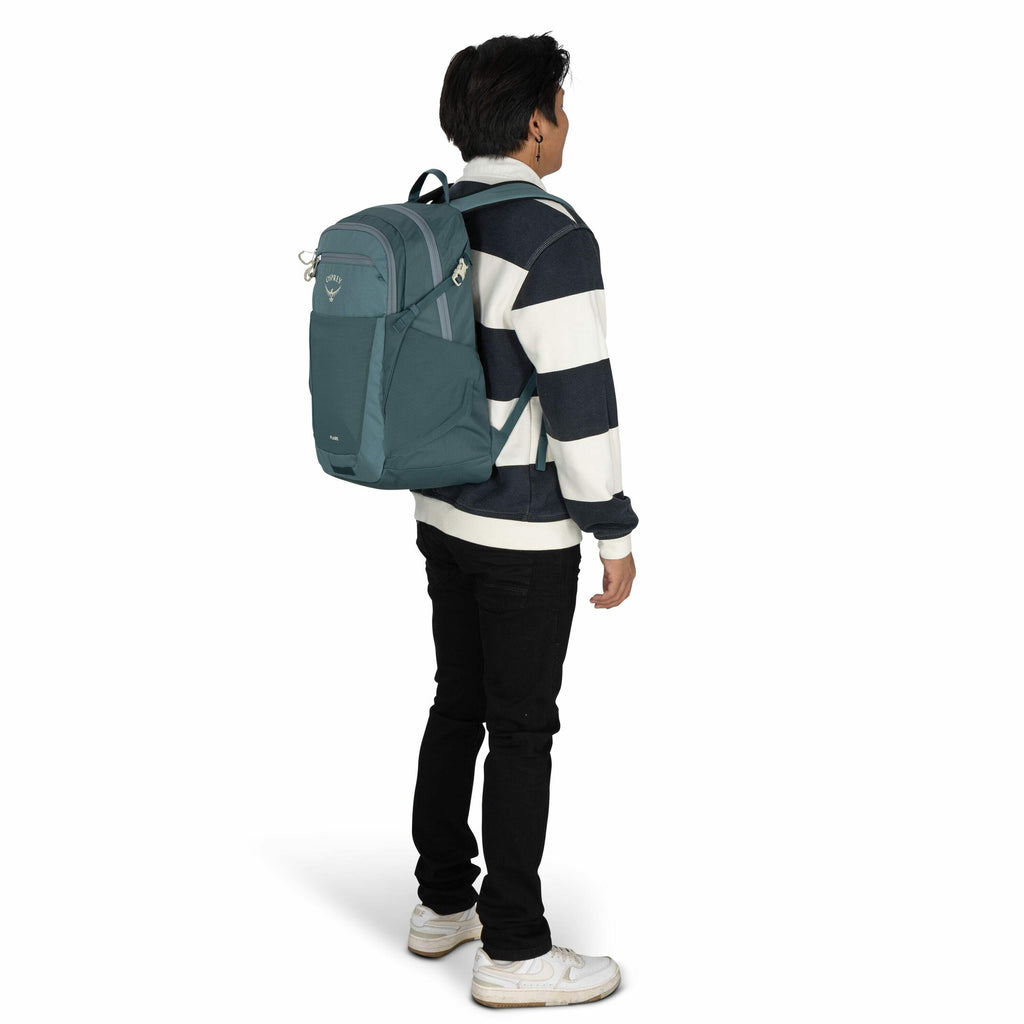 The image shows a person standing and facing slightly to the right, wearing a blue and white horizontally striped sweater, black pants, and white sneakers. The individual is carrying a teal-colored Osprey backpack on their back. The backpack features a lo