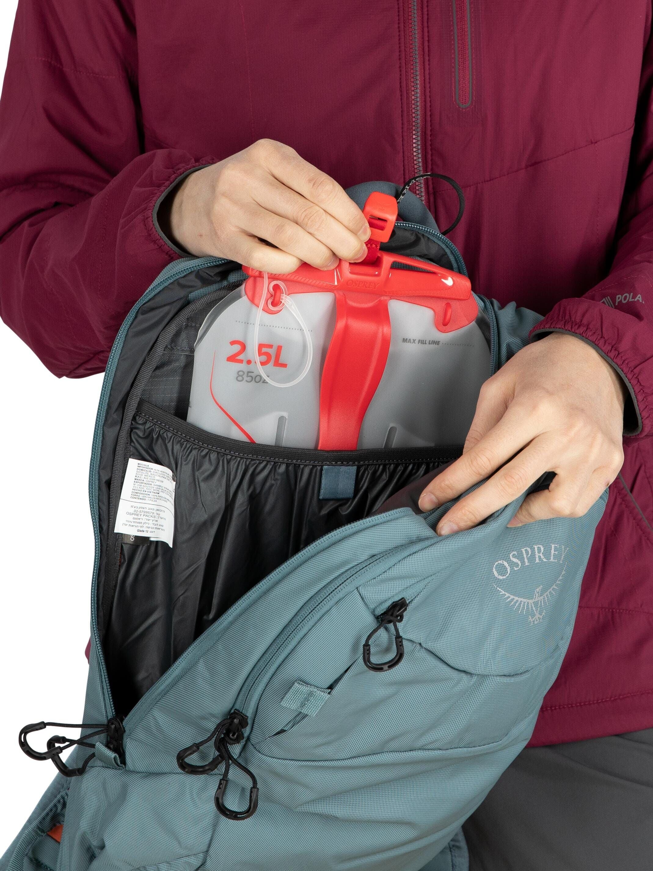 The image shows a person wearing a maroon jacket inserting or removing a hydration reservoir from an Osprey backpack. The reservoir is marked with a capacity of 2.5 liters (85 ounces) and features red accents, including a handle. The person is using both
