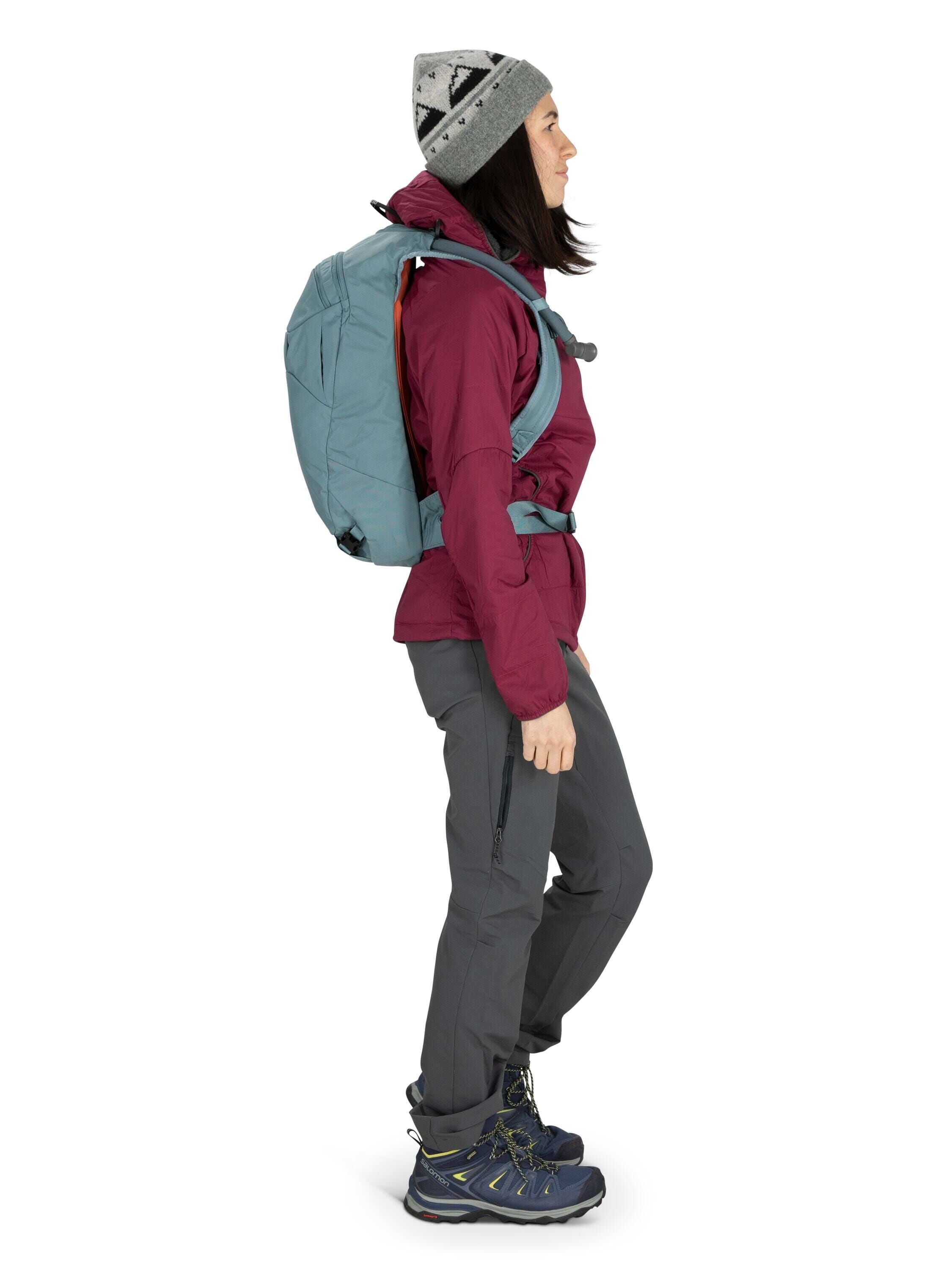 The image shows a person dressed in outdoor hiking attire. They are wearing a maroon jacket and dark gray cargo pants, complemented by a pair of blue hiking boots. A light gray backpack is securely fastened over their shoulders. The individual is also wea