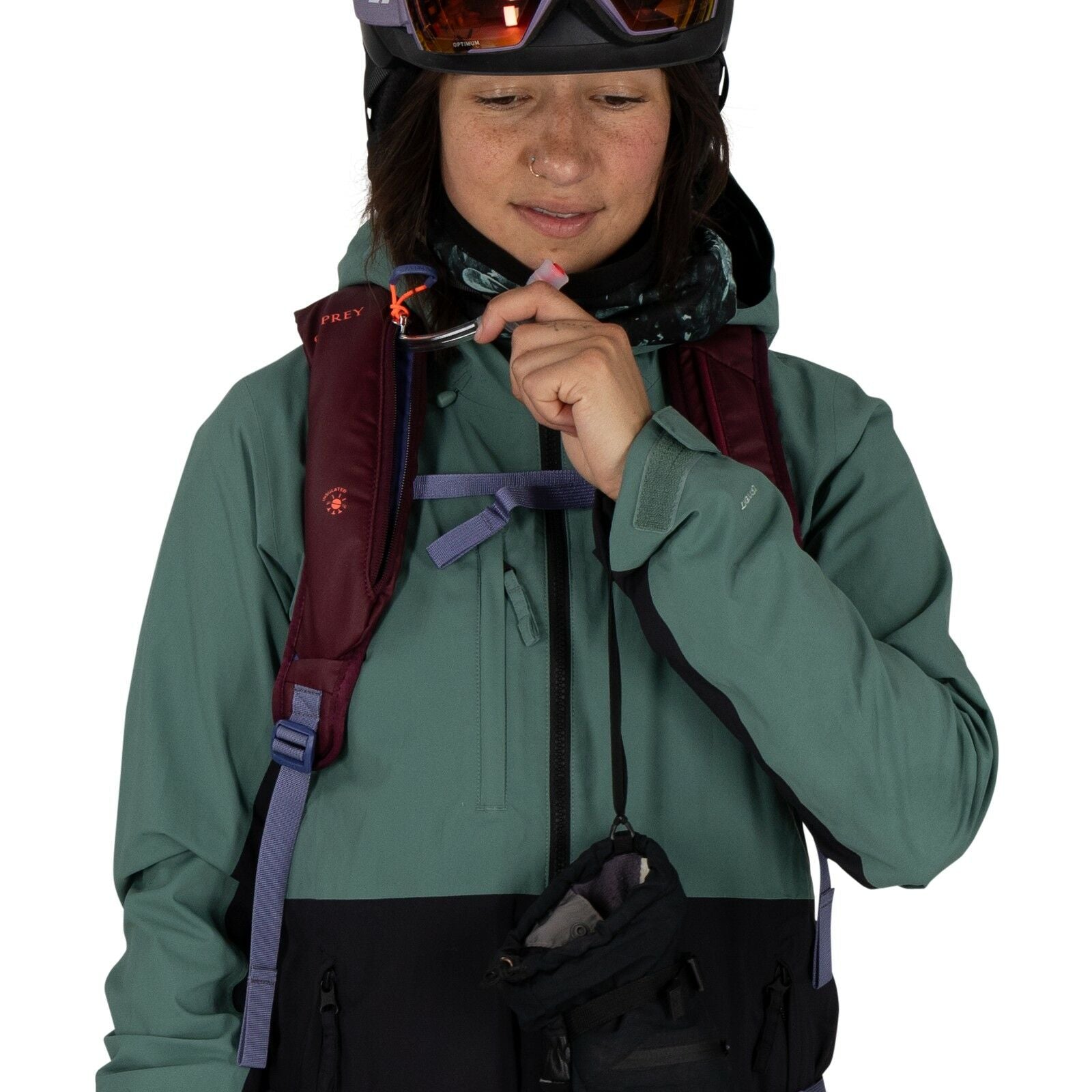 The image shows a person dressed in outdoor or skiing attire, wearing a green and black jacket. Over the jacket, the individual is equipped with a maroon backpack with distinct straps that include a water reservoir tube, which they are holding near their