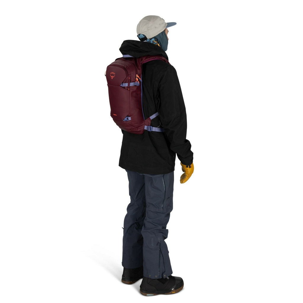 The image shows a person dressed in winter outdoor gear, standing with their side facing the viewer. They are wearing a light gray cap, a blue neck gaiter or scarf covering their lower face, a black jacket, and dark blue pants. The person has on yellow gl