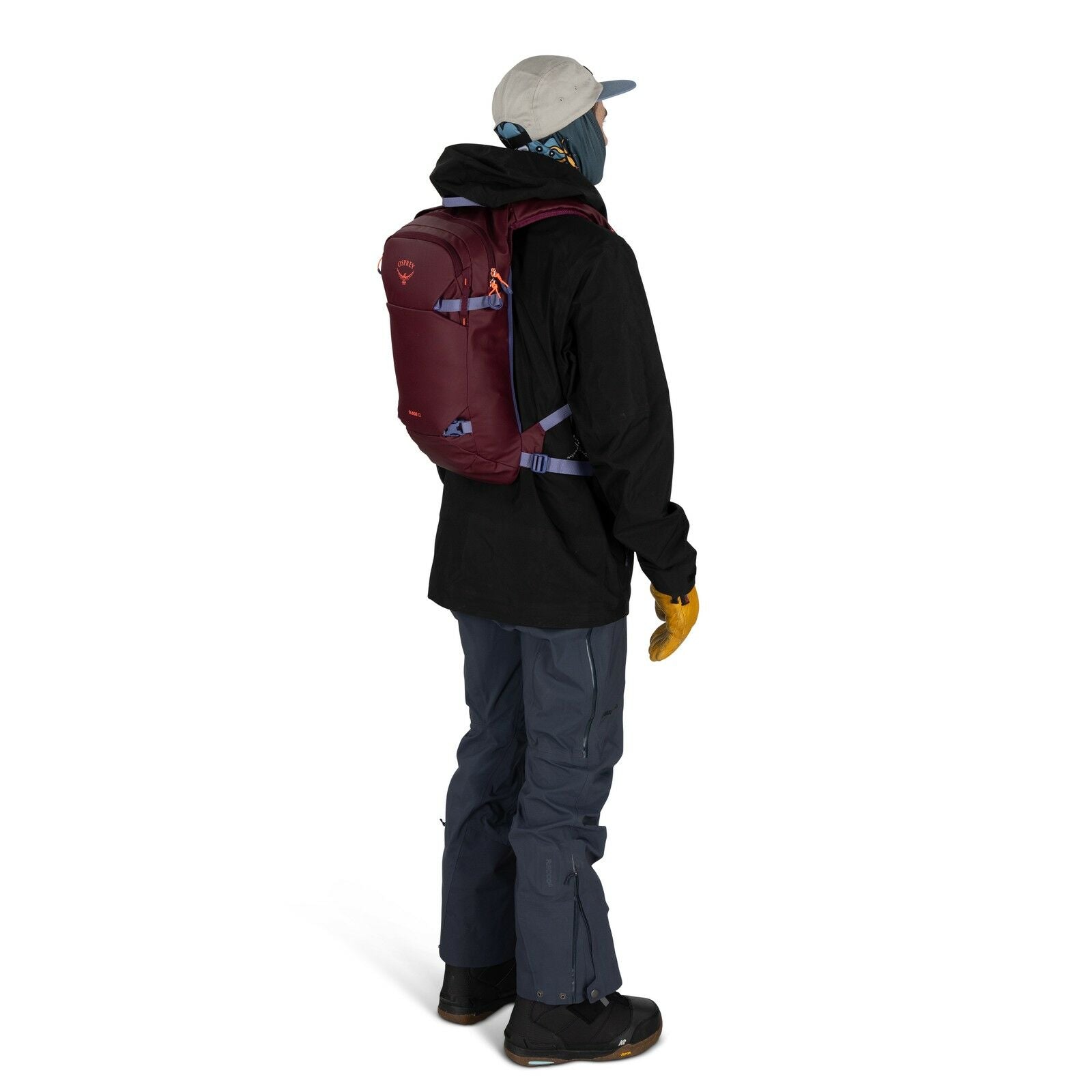 The image shows a person dressed in winter outdoor gear, standing with their side facing the viewer. They are wearing a light gray cap, a blue neck gaiter or scarf covering their lower face, a black jacket, and dark blue pants. The person has on yellow gl