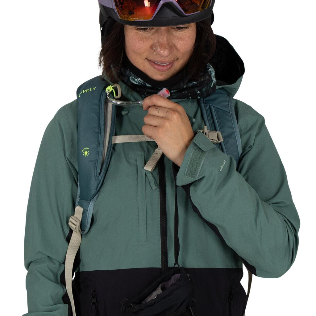 The image depicts an individual wearing outdoor gear, suitable for hiking or other outdoor activities. The person is wearing a teal-colored Osprey backpack, noticeable by its logo on the left shoulder strap. They are dressed in a two-tone jacket with a te