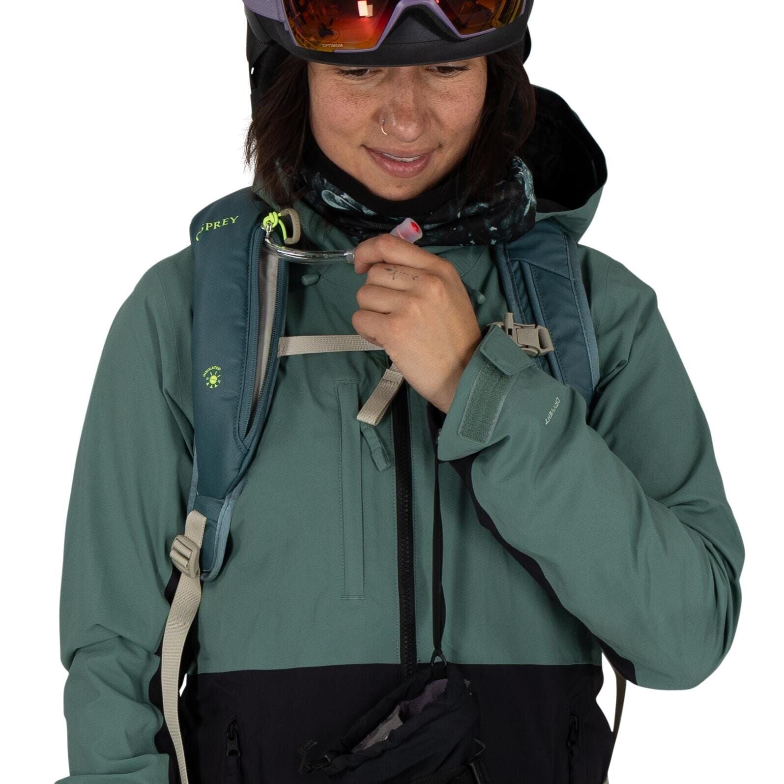 The image depicts an individual wearing outdoor gear, suitable for hiking or other outdoor activities. The person is wearing a teal-colored Osprey backpack, noticeable by its logo on the left shoulder strap. They are dressed in a two-tone jacket with a te