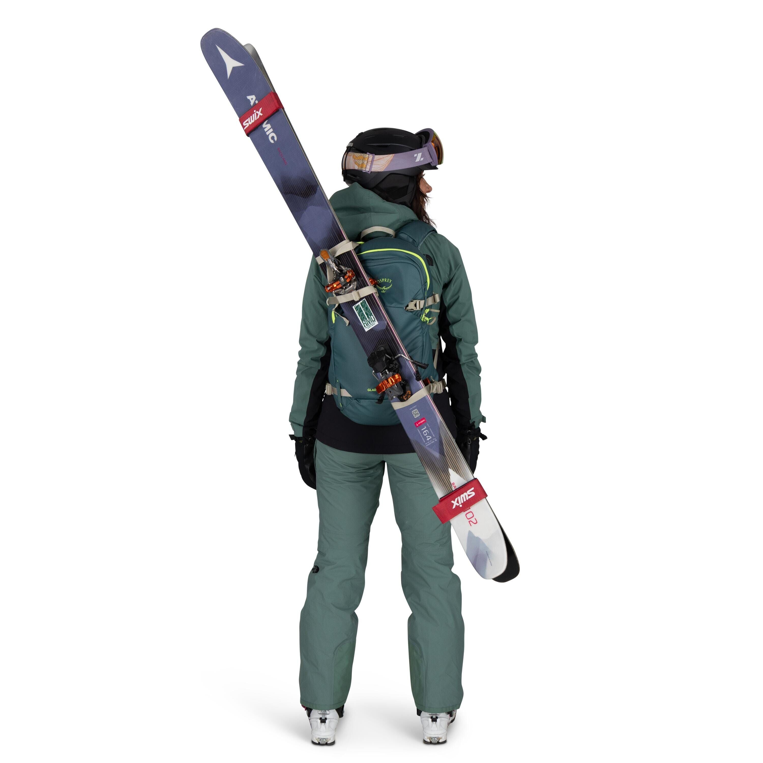 The image shows a person dressed in a complete ski outfit, facing away from the camera. They are wearing a green ski jacket and pants with black gloves and white ski boots. A pair of skis is attached diagonally across their green and blue backpack, which