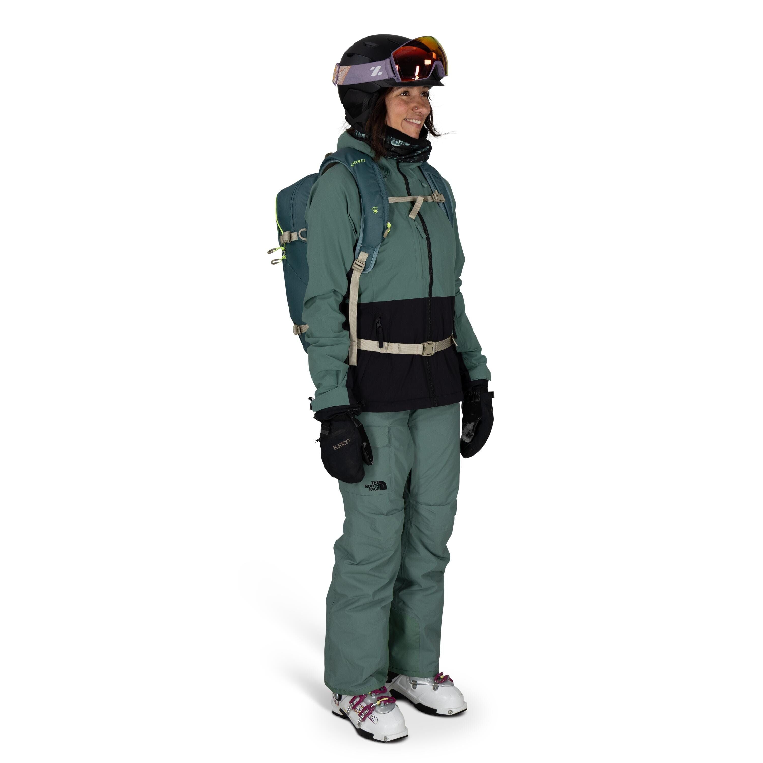 The image shows a person dressed in full ski or snowboarding gear. They are wearing a teal North Face jacket with black accents, matching teal snow pants, and white ski boots with red and black detailing. The person is equipped with a black ski helmet and