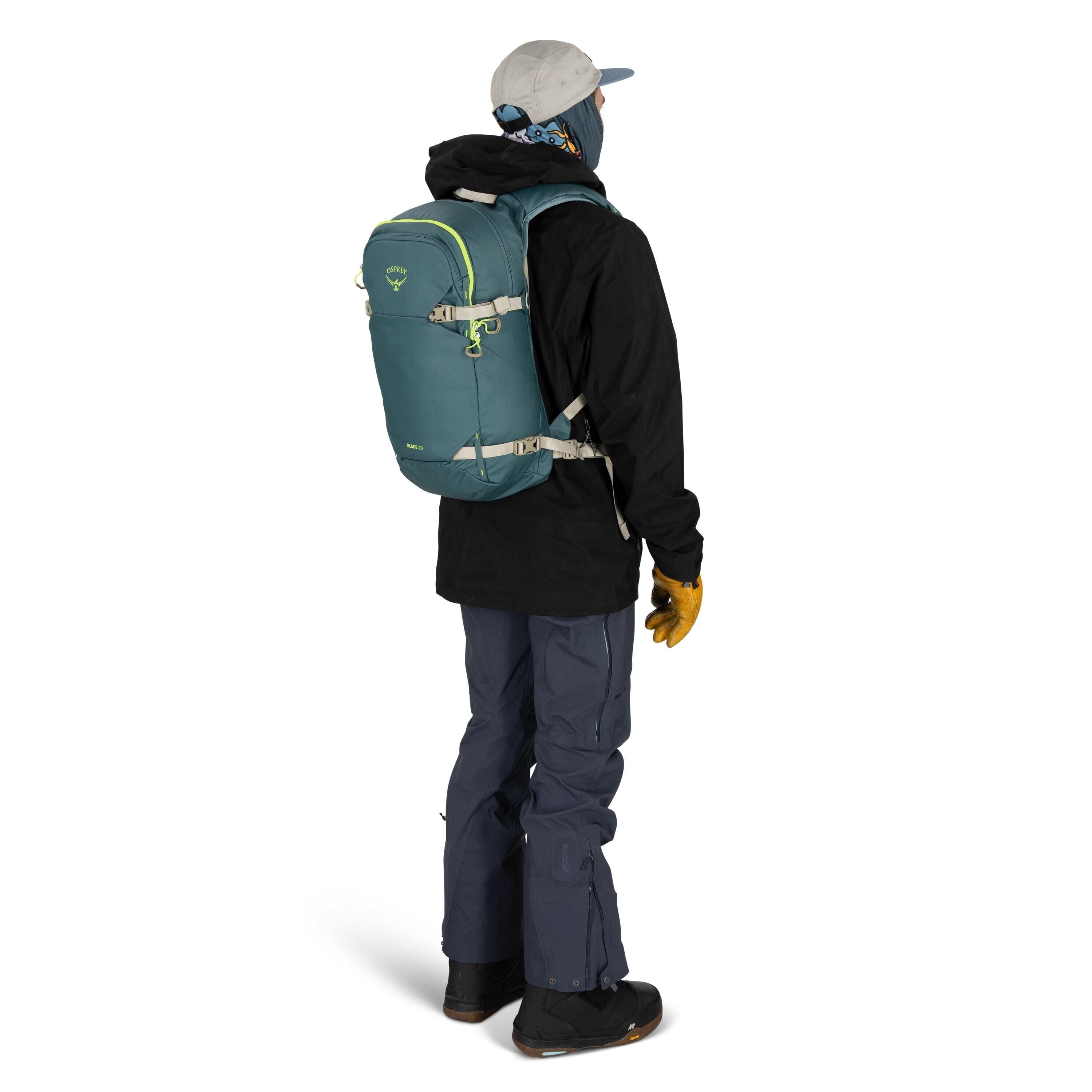 The image depicts a person standing and facing away from the camera, dressed in outdoor winter apparel. They are wearing a light gray cap, a dark neck gaiter, and a black jacket, which covers most of their upper body. The person has on blue cargo pants an