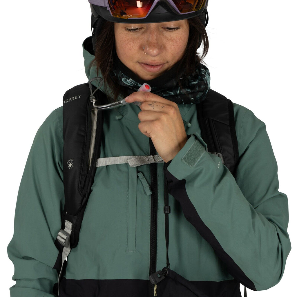 The image shows a person wearing a green and black outdoor jacket, suitable for cold weather or snow sports. They have a black Osprey backpack, which suggests they might be preparing for an outdoor activity such as hiking or skiing. The individual is hold