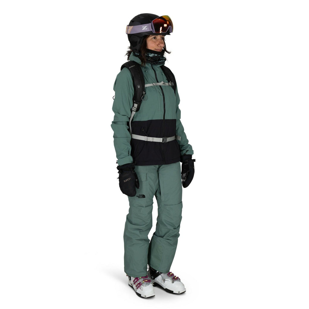 The image shows a person dressed in outdoor winter gear suitable for skiing or snowboarding. They are wearing a green and black The North Face jacket paired with matching green pants and black gloves. The outfit is accessorized with a backpack, and the pe