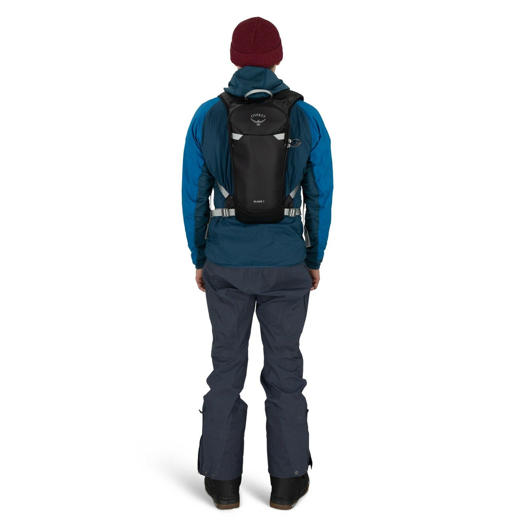 The image shows a person standing facing away from the camera, wearing outdoor gear suitable for cold weather. They are dressed in a blue jacket and dark pants, with black boots on their feet. A maroon beanie covers their head, and they have a black backp