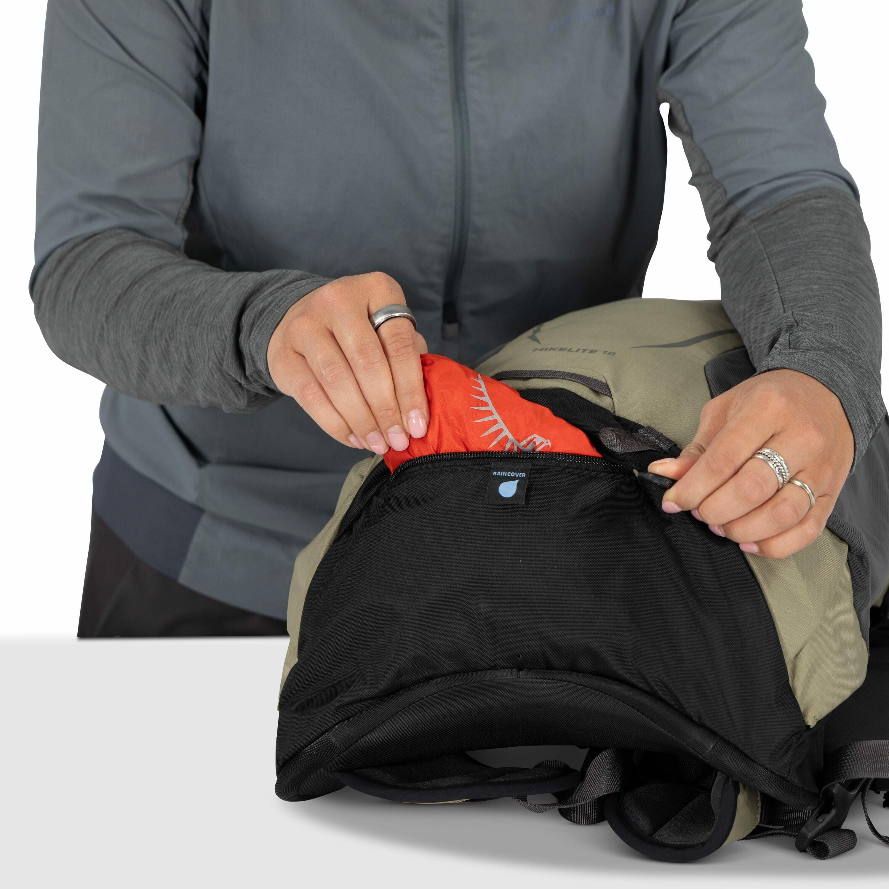 The image shows a person dressed in a gray jacket, who is unzipping a pocket on a backpack. Inside the pocket, there is a bright red rain cover with a white logo resembling a stylized feather or wing design. The backpack appears to be a light green color