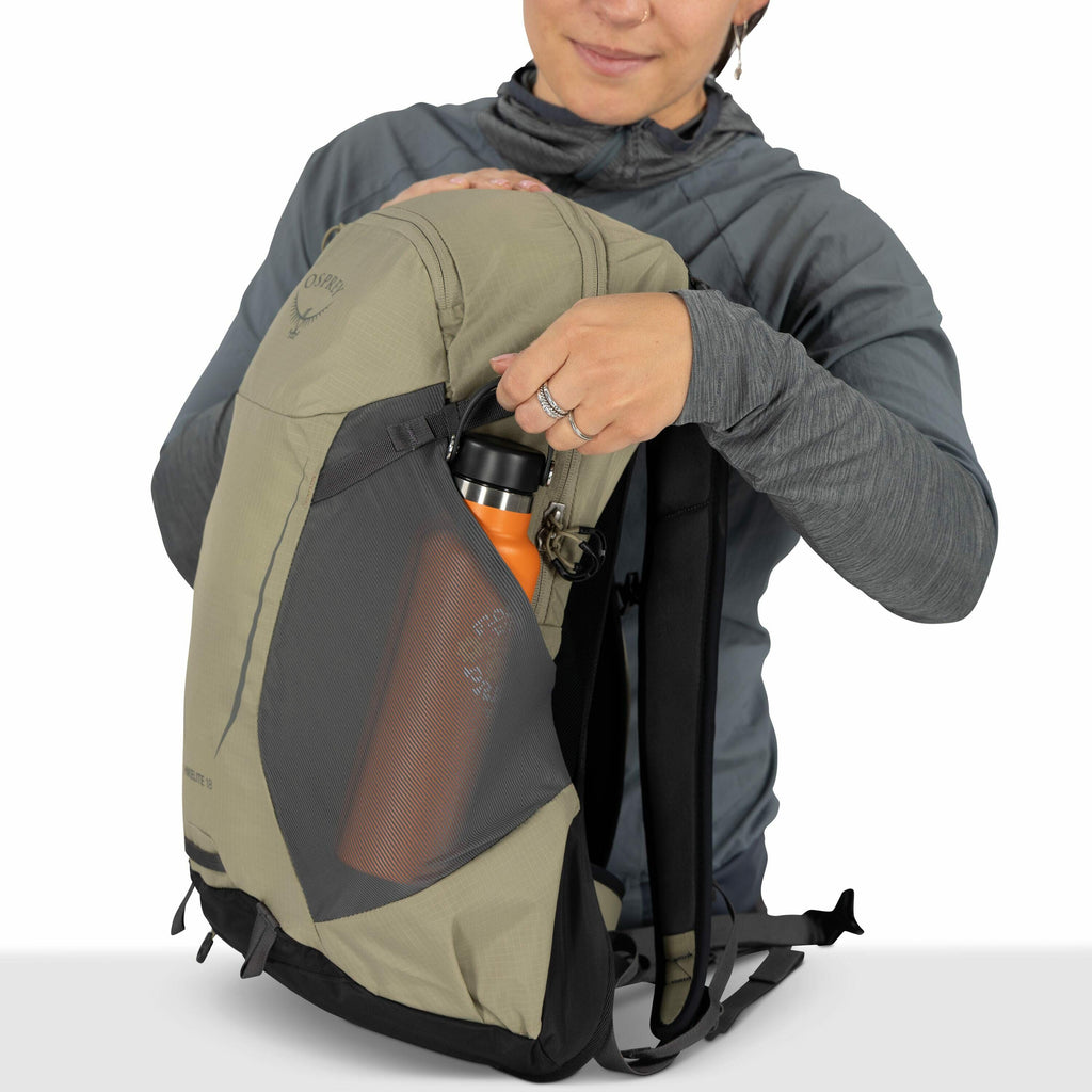 The image shows a person holding a beige and black backpack, revealing a bright orange water bottle stored in the mesh pocket on the side. The backpack features a visible Osprey logo near the top. The person is wearing a gray, long-sleeved jacket with a h