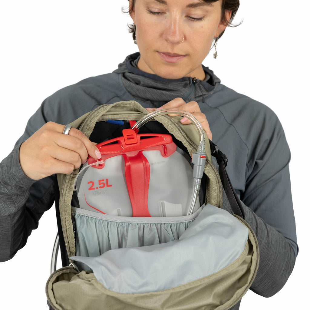 The image shows a person holding a backpack with its main compartment open to reveal the contents. Inside, there is a hydration reservoir with a red cap and a tube, designed for convenient drinking during activities like hiking. The reservoir is labeled w