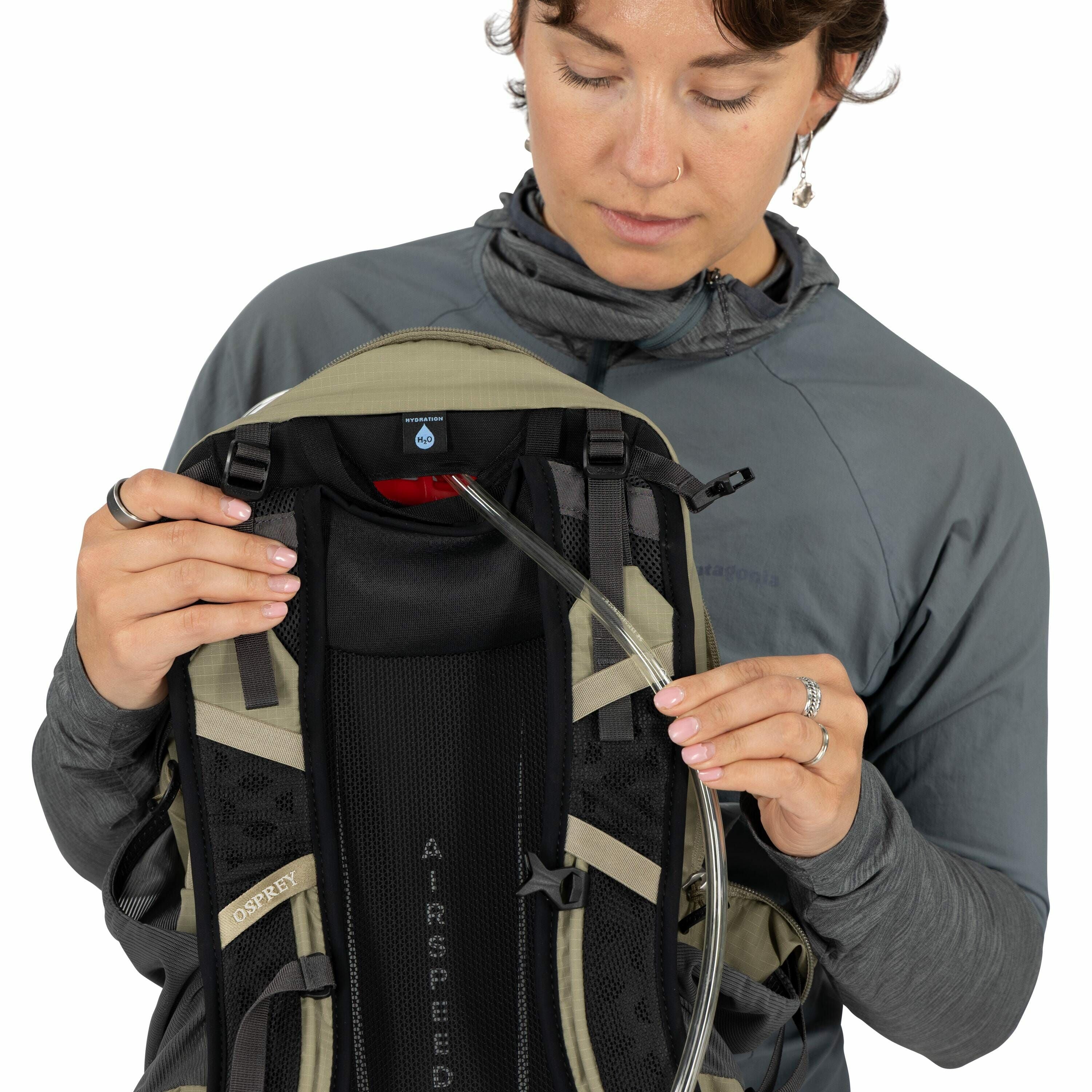 The image shows a person wearing a Patagonia jacket and holding an Osprey backpack. The backpack features the word "AIRSPEED" on its back panel and includes a hydration system with a visible hose extending from a compartment at the top. The compartment is