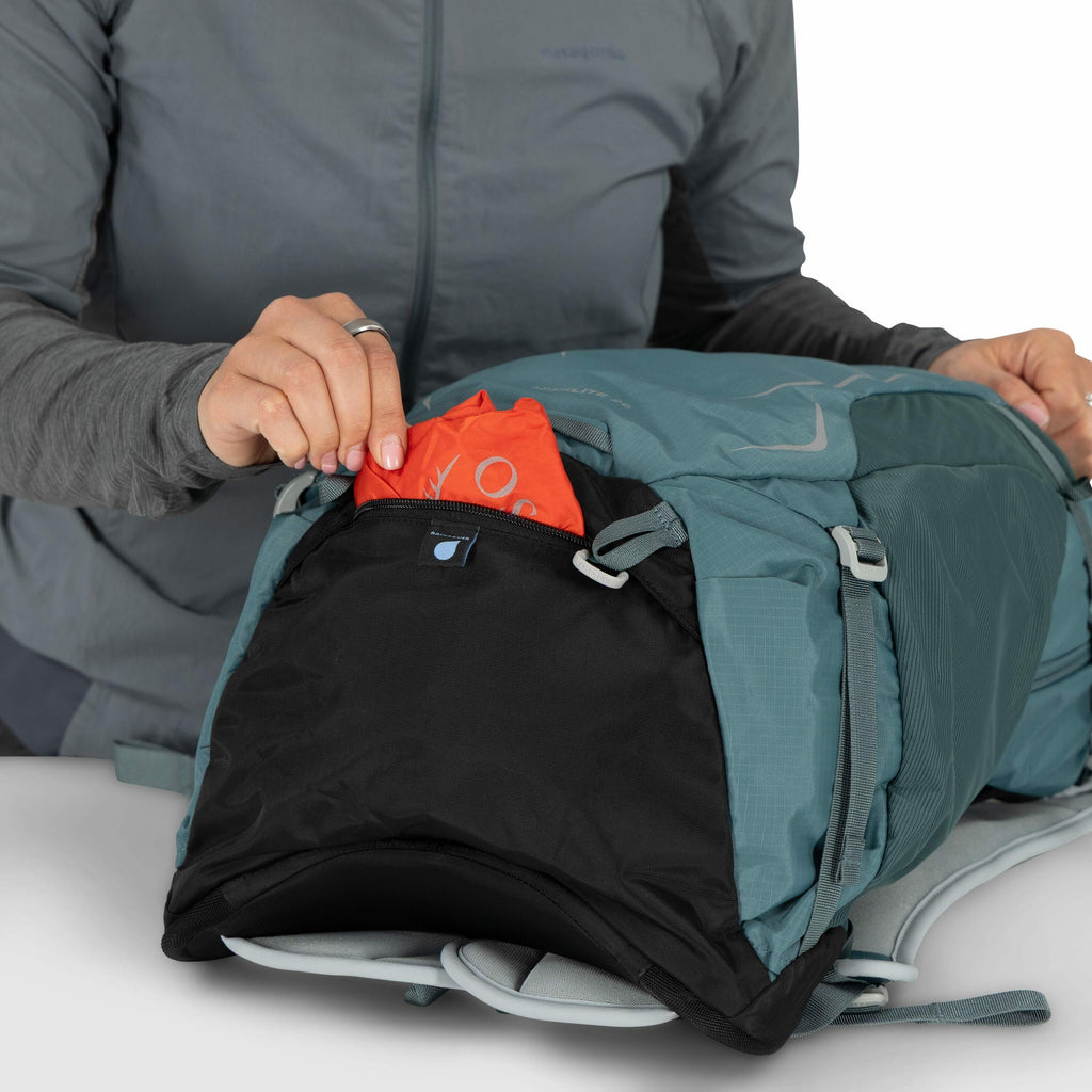The image depicts a person wearing a grey Patagonia jacket, placing an item into the front pocket of a teal backpack. The backpack is made of durable material with light grey accents, straps, and buckles. The item being stored appears to be a vibrant oran