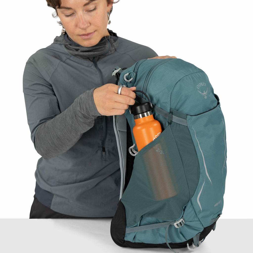 The image shows a person wearing a grey jacket over a black shirt, holding a blue Osprey backpack. The person is placing or retrieving an orange water bottle from a mesh side pocket of the backpack. The Osprey logo is visible on the top of the backpack, w