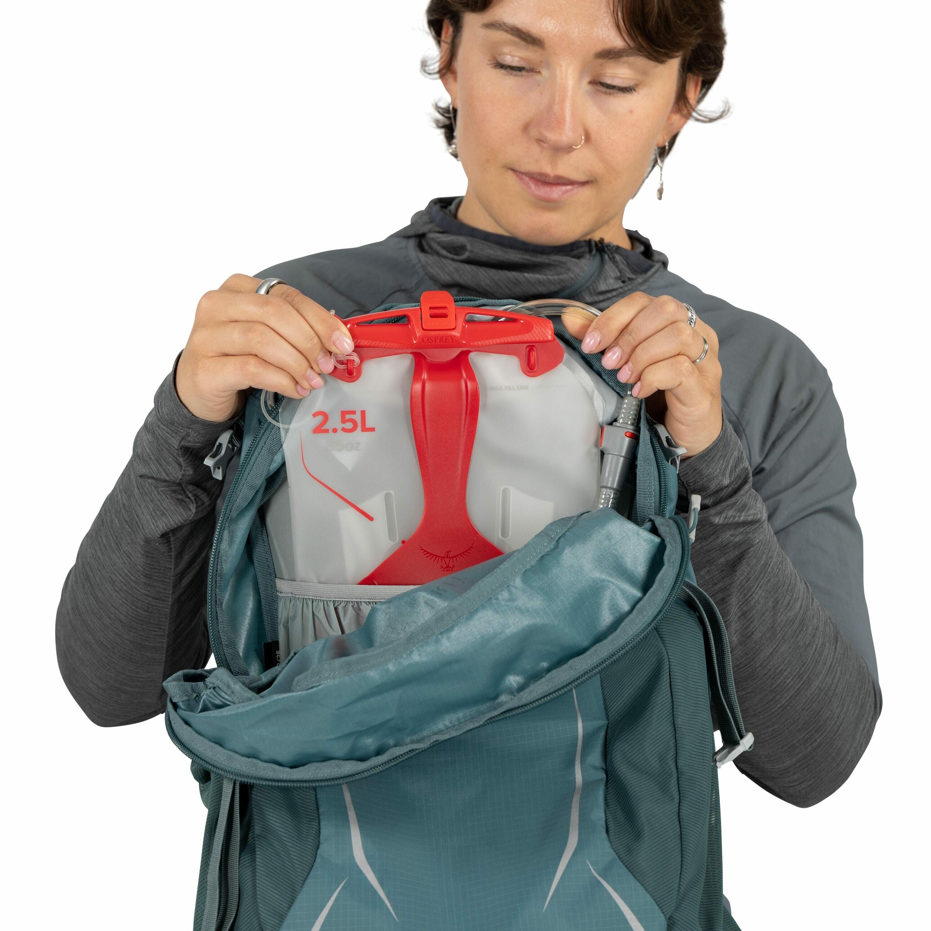 The image depicts a person holding open a teal-colored backpack to reveal a hydration reservoir inside. The reservoir is labeled with a capacity of "2.5L" and has a distinctive red handle and accents, indicating it is designed for easy carrying and use. T