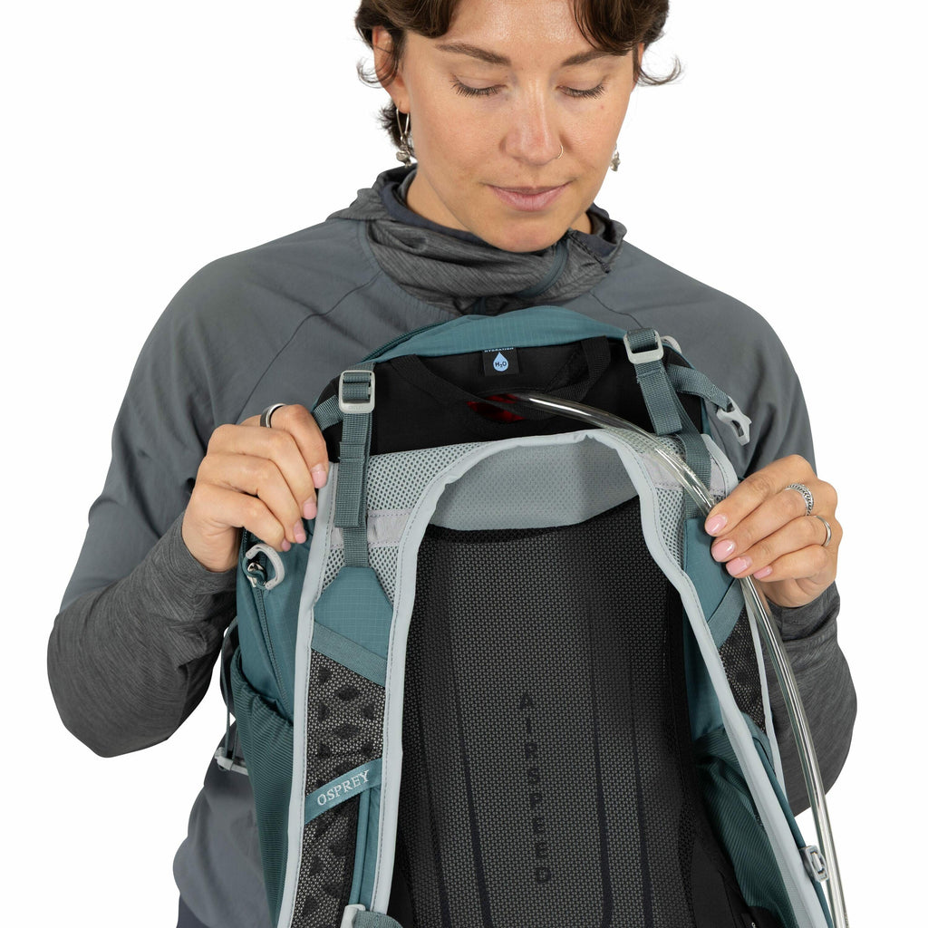 The image shows a person wearing a grey long-sleeve shirt while holding an Osprey backpack. The backpack is a teal color with a black and grey Airspeed mesh back panel designed for ventilation and comfort. A hydration tube runs over the top of the backpac