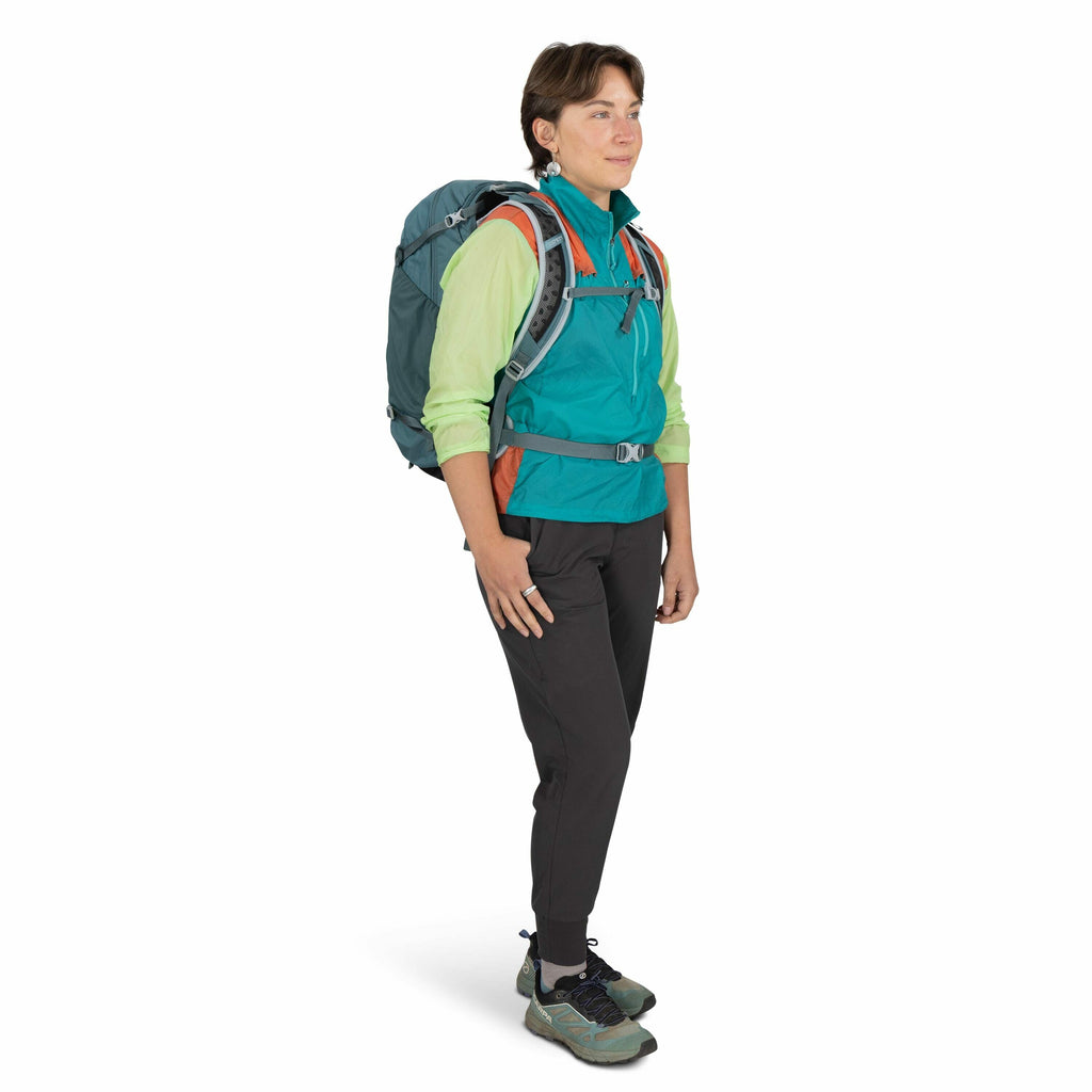 The image shows an individual wearing outdoor hiking attire, including a turquoise and orange jacket layered over a light green shirt. The person is also wearing dark gray pants and gray and teal hiking shoes. A large, dark teal hiking backpack is strappe