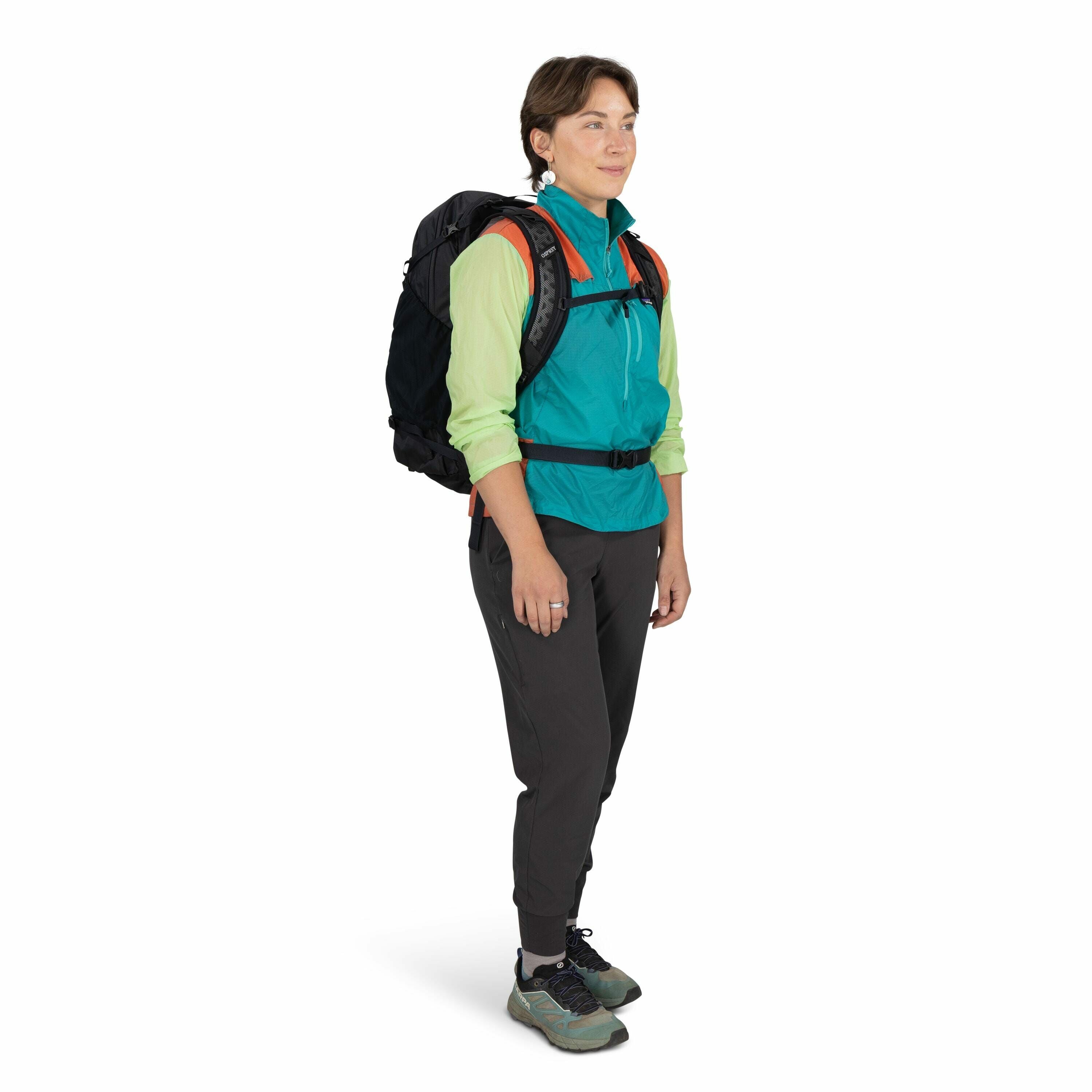 The image shows a person standing in a white studio background, dressed in casual outdoor attire suitable for hiking or trekking. They are wearing a vibrant outfit consisting of a turquoise jacket with a collar and rolled-up sleeves layered over a long-sl