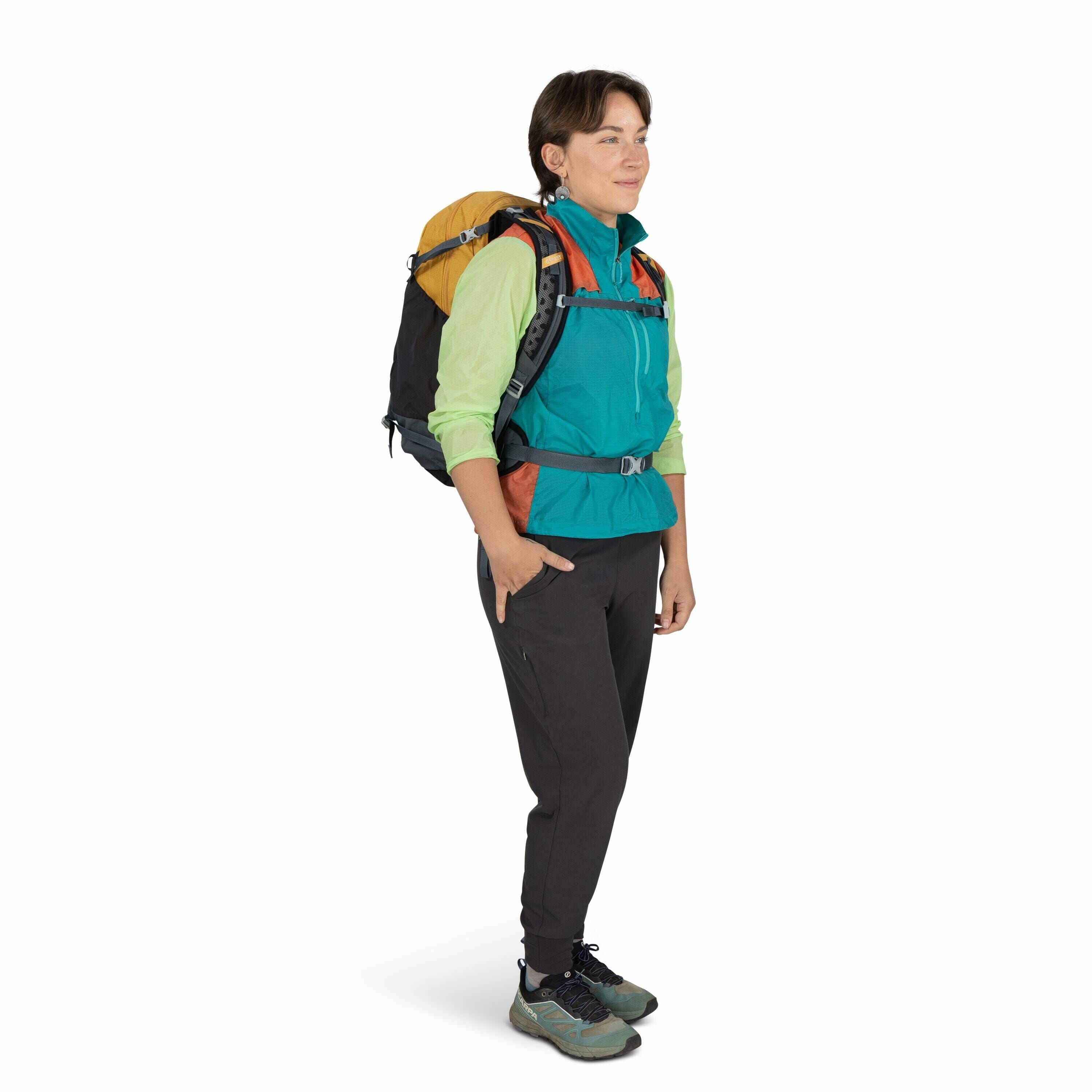 The image shows a person standing and dressed for a hiking trip. They are wearing a vibrant outfit that includes a teal and green jacket with the sleeves rolled up and black hiking pants. On their feet are sturdy green and black hiking shoes. The individu