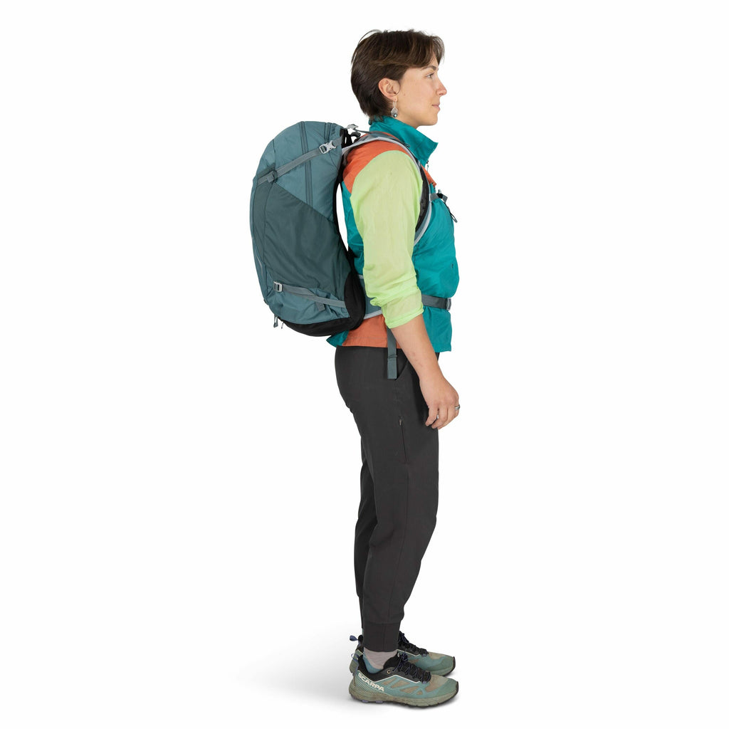 The image shows a person standing in a profile view, dressed in outdoor attire, and ready for an adventure. They are wearing a vibrant, multi-colored outfit that includes a blue and orange vest over a green long-sleeve shirt, paired with dark, fitted pant
