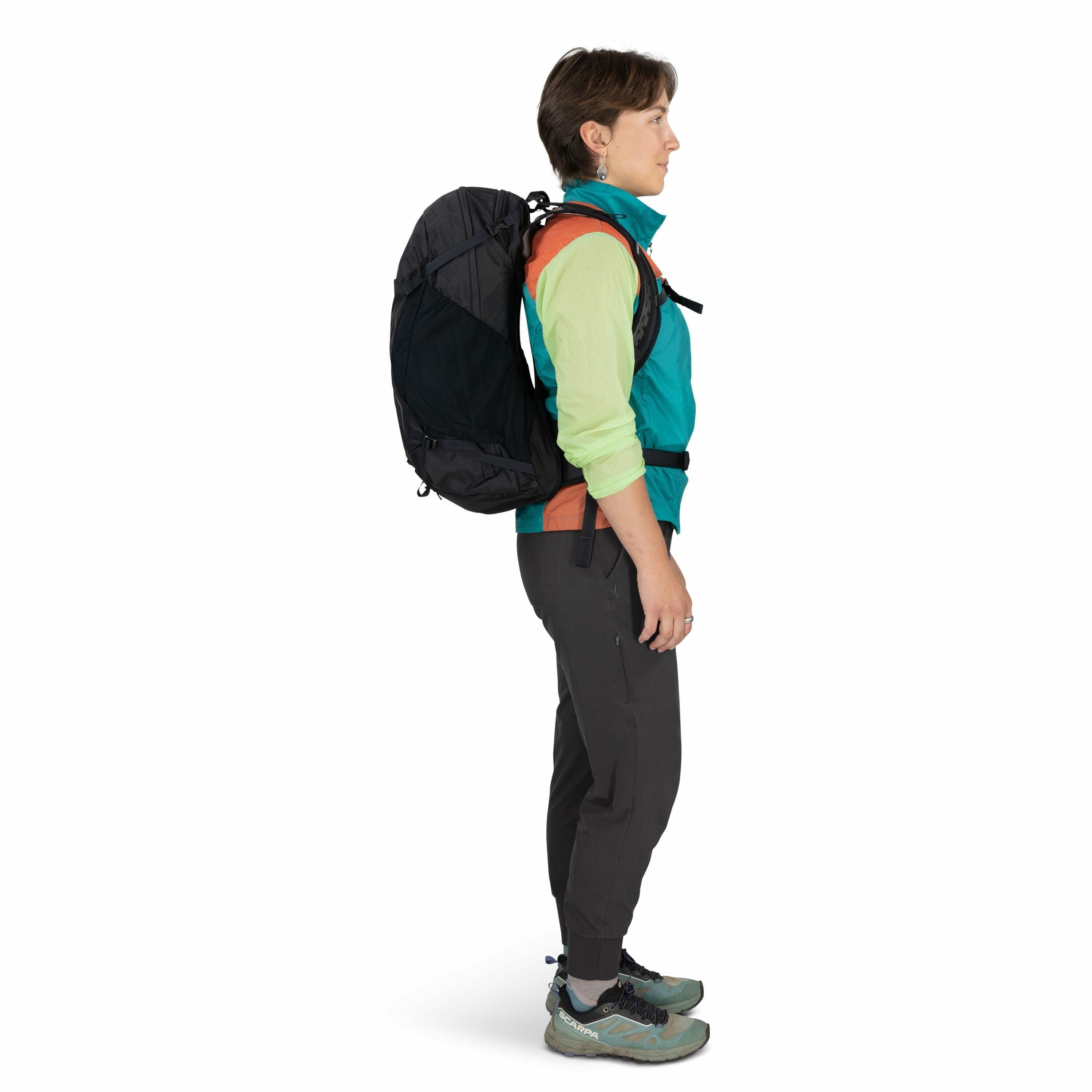 The image depicts a person in a side profile, standing against a plain white background. The individual is dressed in outdoor gear, including a multicolored jacket with teal, orange, and lime-green sections, and dark gray hiking pants. They are wearing a