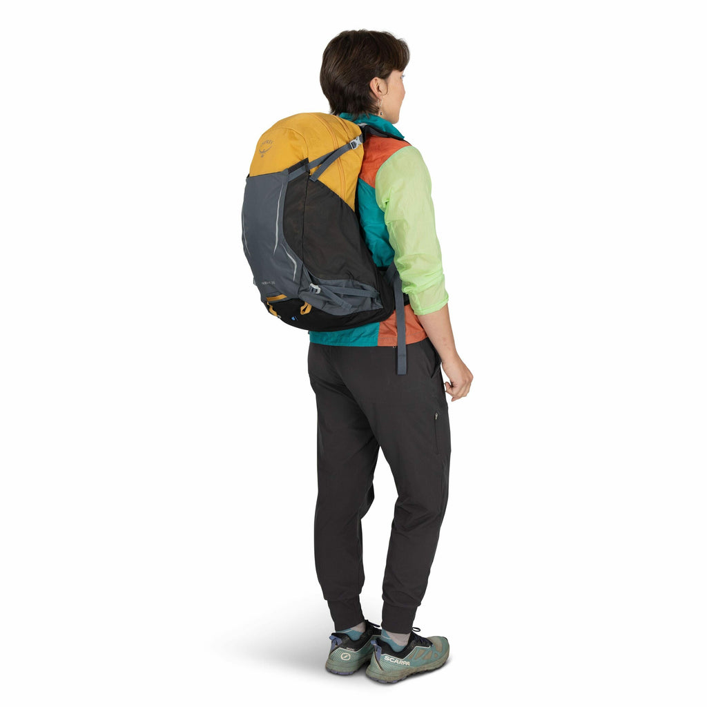 The image features a person wearing outdoor attire, viewed from the back. The individual is wearing a multicolored jacket with panels of teal, red-orange, and light green and dark-colored pants. They have a large backpack on their back, which is primarily