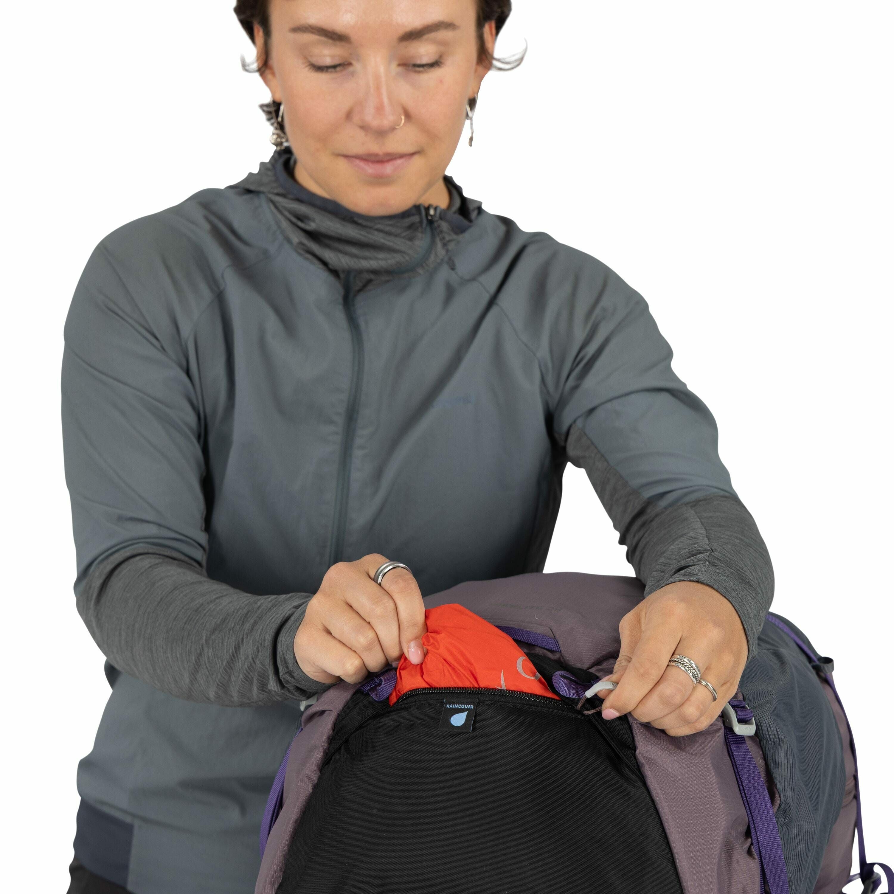 The image shows a person wearing a long-sleeved gray jacket over a darker gray top, standing in front of a backpack. The person is in the process of closing a black compartment on the backpack, using two zippers. Inside the backpack, a bright red item is