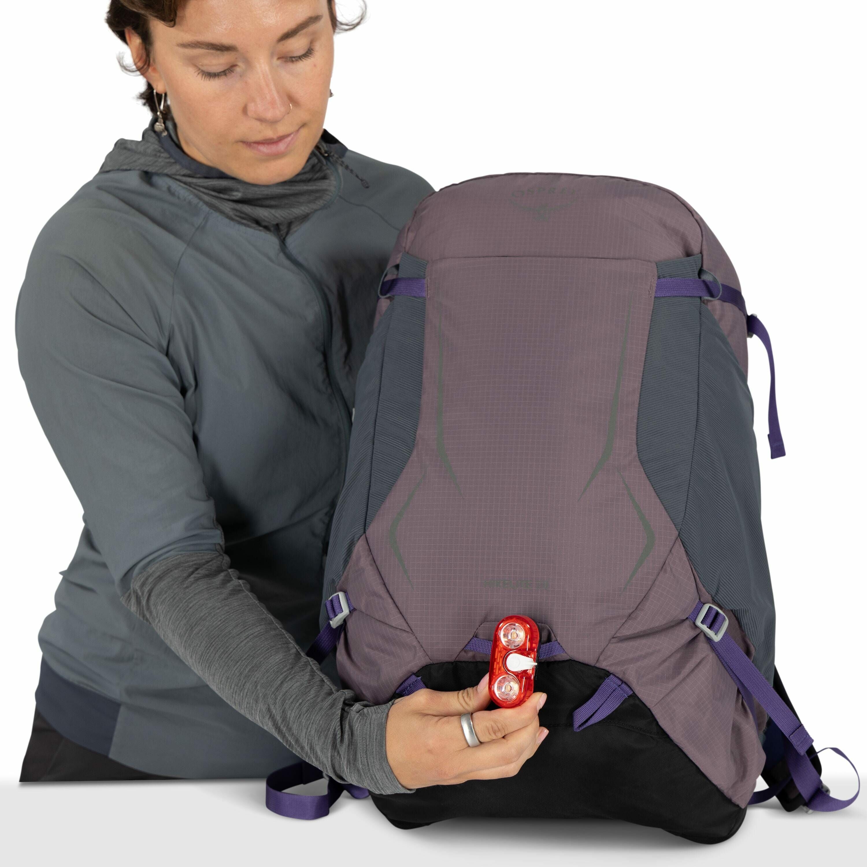 The image depicts an individual holding a large Osprey backpack. The backpack is predominantly purple with gray accents and features purple straps for adjustments. The person is attaching or holding a red light with two bulbs at the bottom of the backpack