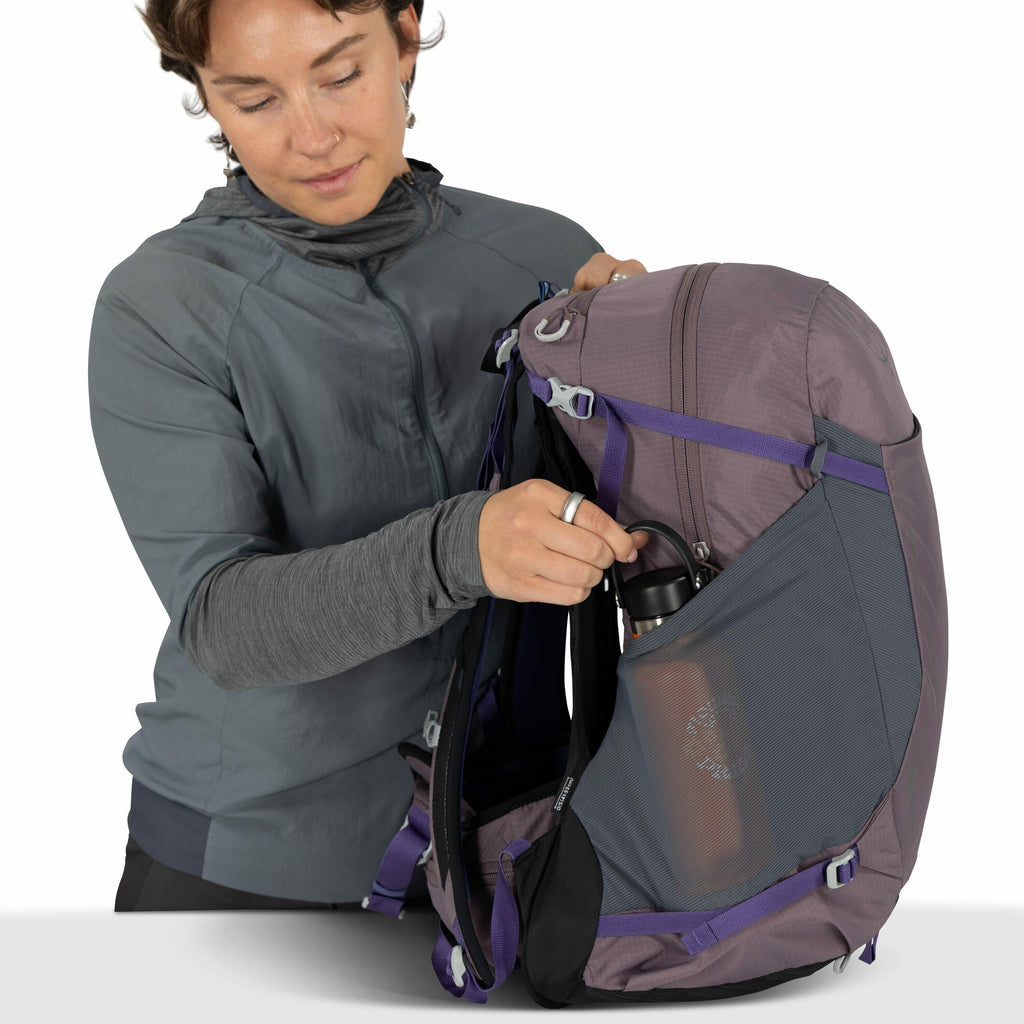 The image features a person holding a large outdoor backpack. The backpack is predominantly gray with purple straps and accents, designed with multiple compartments and external mesh pockets for storage and accessibility. The person is placing a black wat