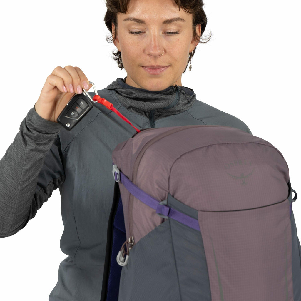 The image shows a person wearing a dark grey, long-sleeve jacket. They are holding a set of car keys attached to a red strap which is connected to a carabiner hooked onto a purple backpack. The backpack has the Osprey logo visible on the top section. The