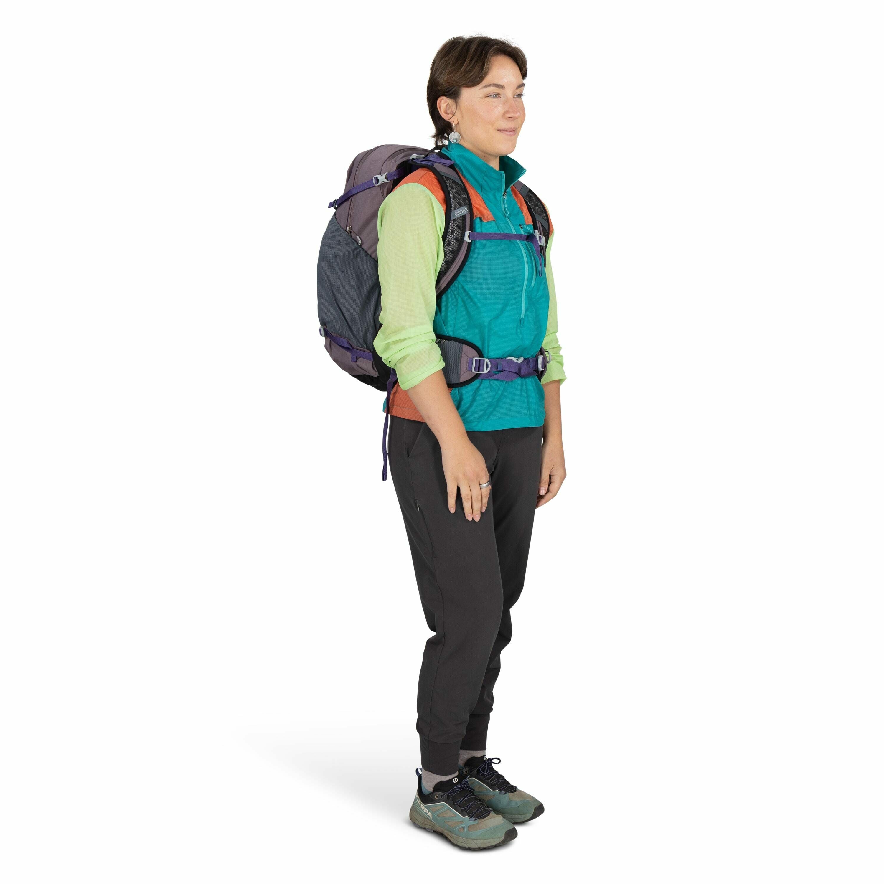 The image shows a person standing in a side profile, wearing hiking gear. The individual is equipped with a large, dark-colored backpack secured with blue and purple straps. They are dressed in a vibrant turquoise and green long-sleeve jacket layered over