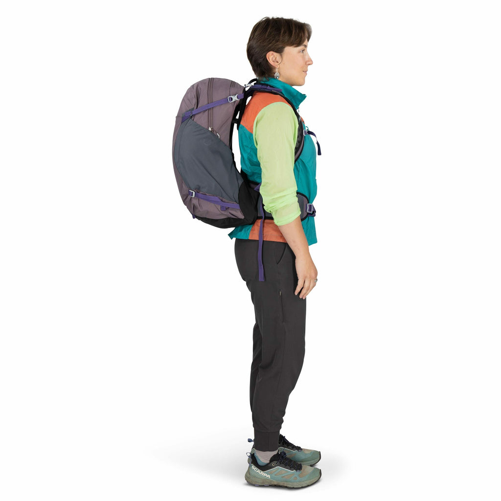 The image shows a person standing in profile view, equipped for hiking. They are wearing a colorful hiking outfit consisting of an orange top layered with a turquoise and green zip-up jacket. Their hiking pants are dark gray, complemented by a pair of tea