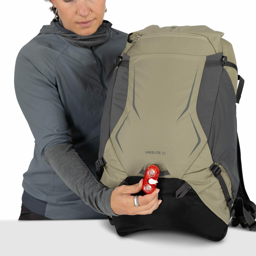 The image depicts a person holding a large hiking backpack branded as "HIKELITE 32." The backpack is primarily gray with some dark accents and has a streamlined design suitable for outdoor activities. The person is wearing a dark gray, lightweight jacket