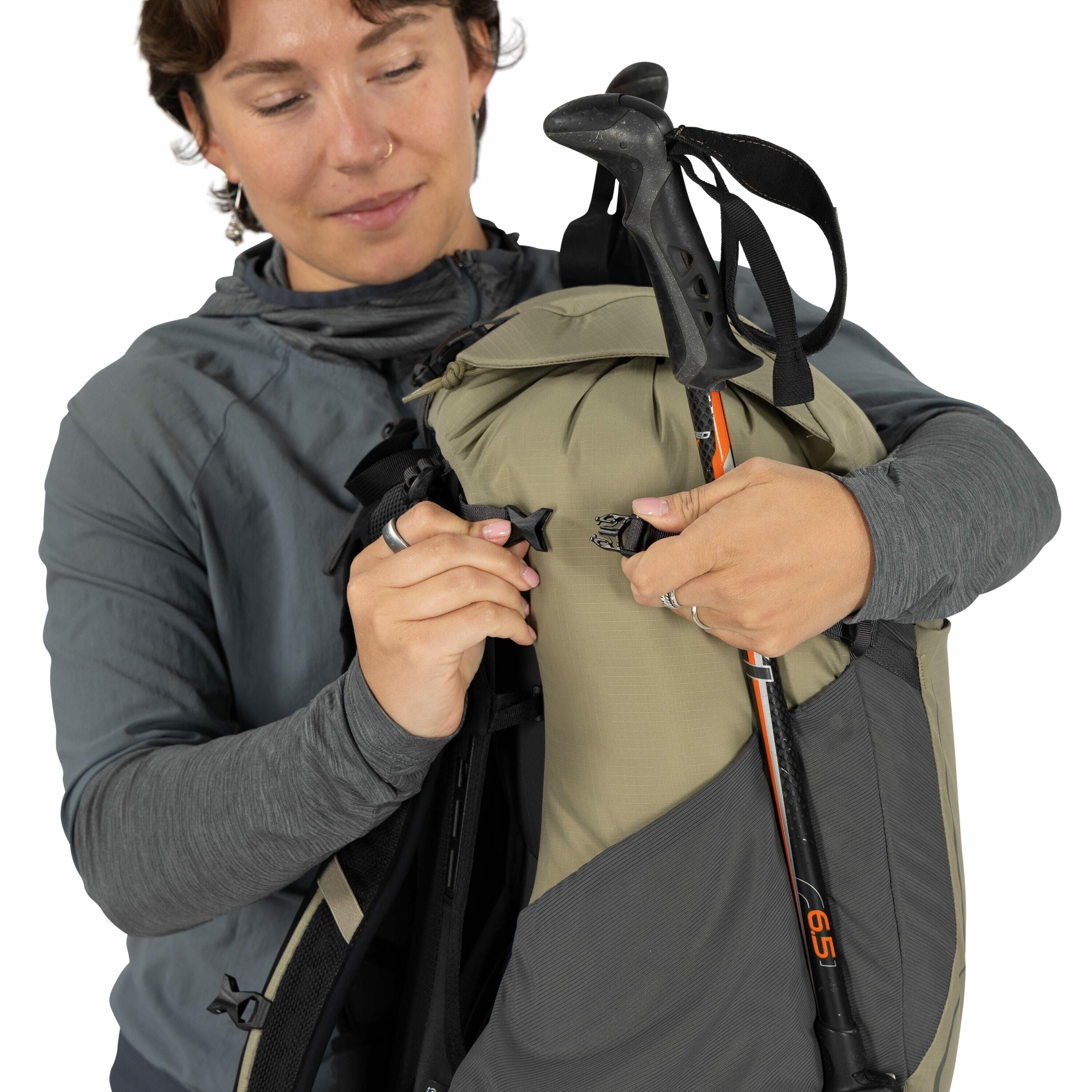 The image shows a person wearing a long-sleeved, grey outdoor jacket while adjusting a light brown and grey backpack. The backpack features a trekking pole securely attached to its side with an orange marking indicating "65," suggesting it might have a 65