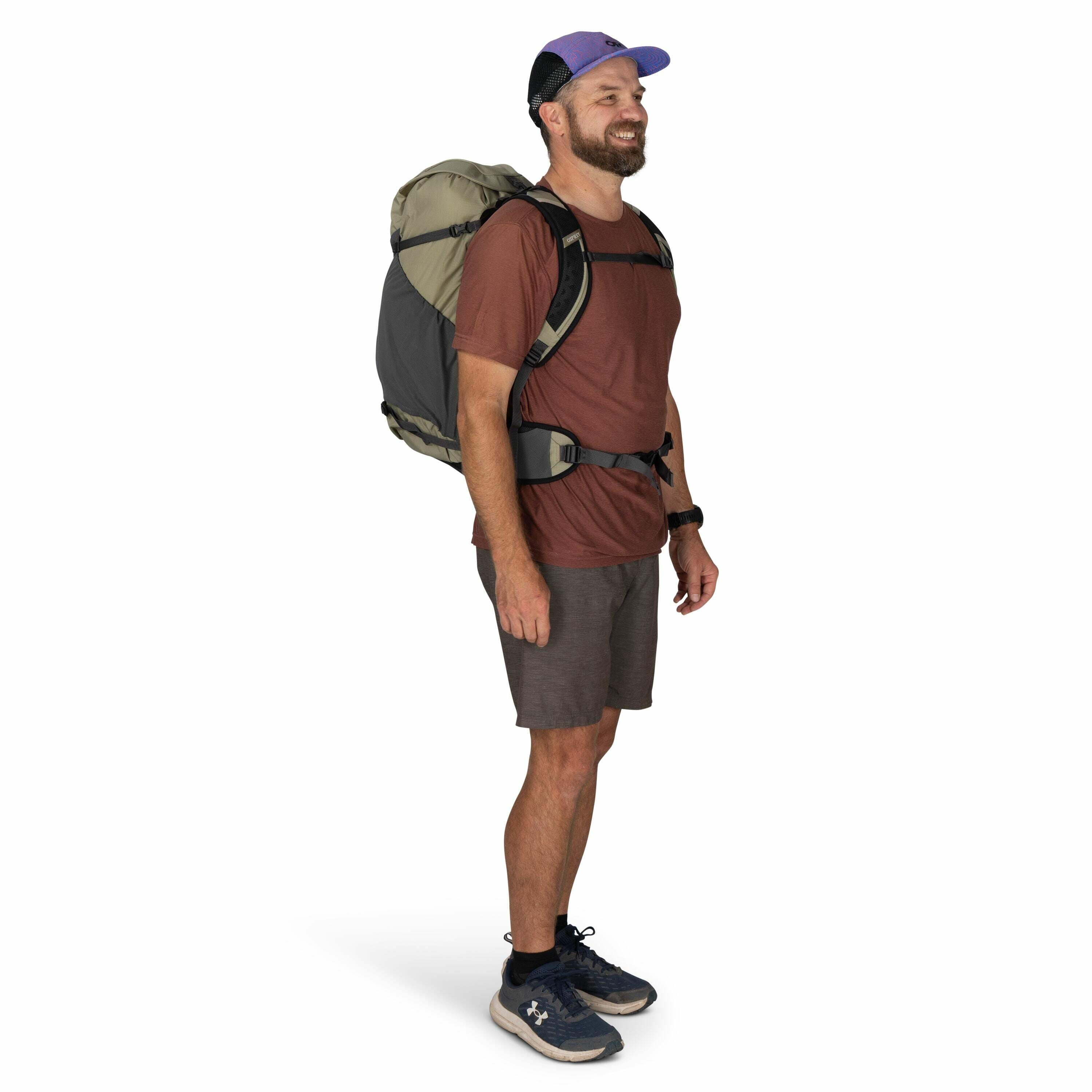 The image shows a person standing in a studio setting wearing casual outdoor attire. They are dressed in a reddish-brown t-shirt paired with dark gray shorts and navy blue athletic shoes. The person is carrying a large backpack, which is primarily green a