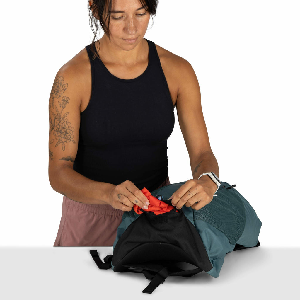 The image shows a person standing at a table, packing a teal-colored backpack. The individual is wearing a black sleeveless top and pink shorts, with tattoos visible on their left arm, including floral designs and a butterfly. Their fingernails have a lig
