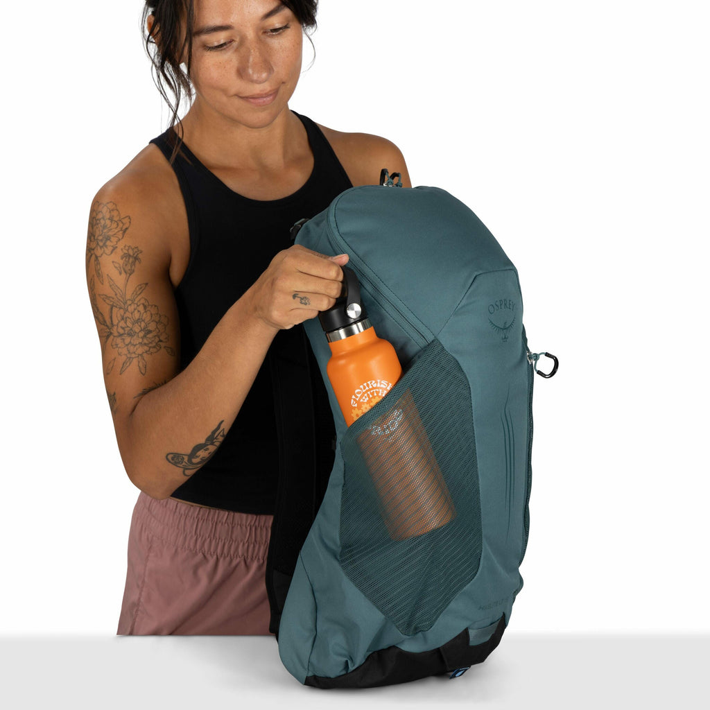 The image shows a person holding a backpack with one hand while placing a bright orange water bottle into the mesh side pocket with the other. The backpack is an Osprey brand, evident from the logo visible on its front. The individual is wearing a sleevel